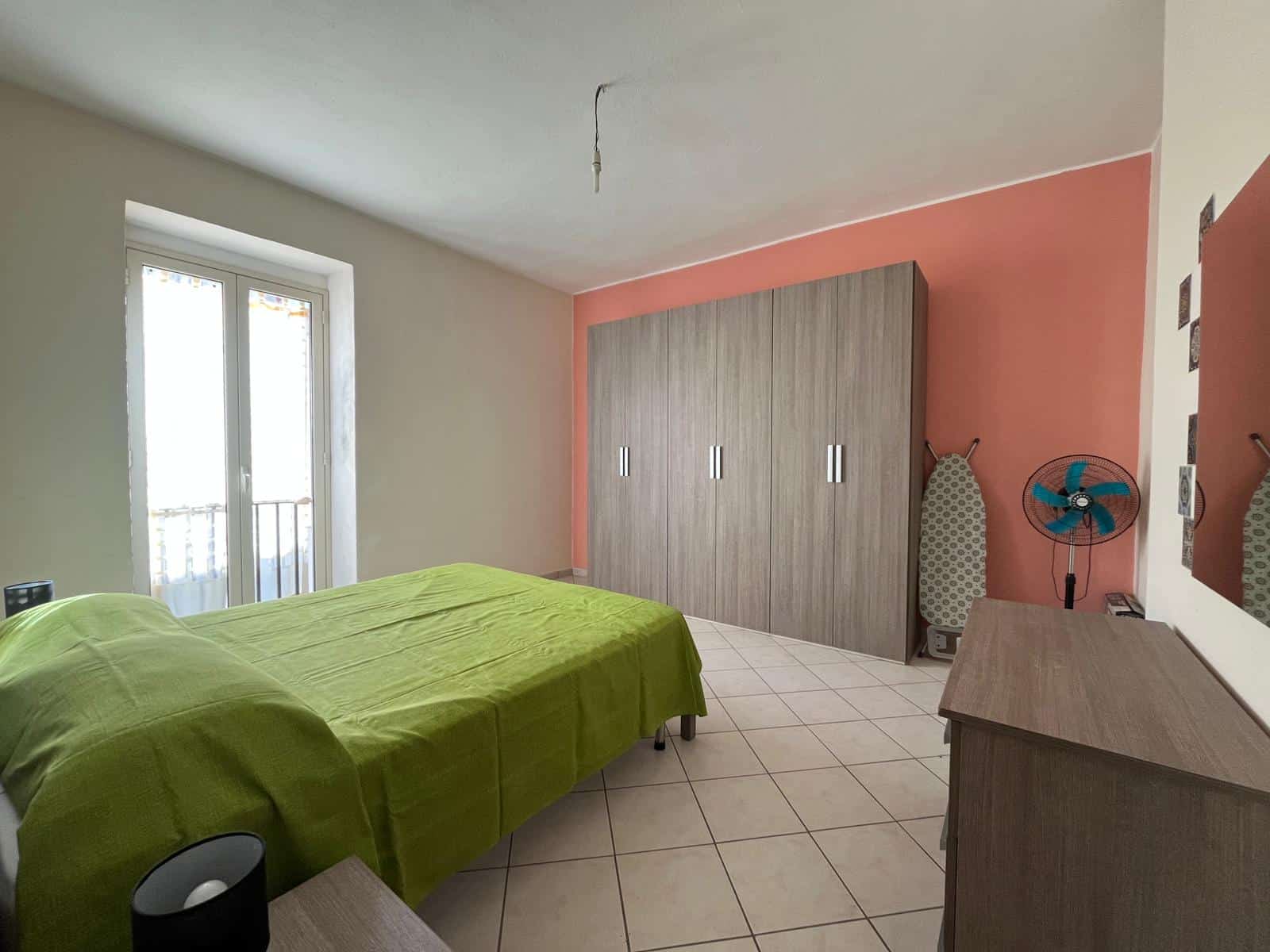 Una camera da letto moderna con pareti bianche e una parete accentuata rosa, arredi in legno chiaro, un armadio spazioso e un letto con copertura verde, illuminata da luce naturale attraverso una fine.