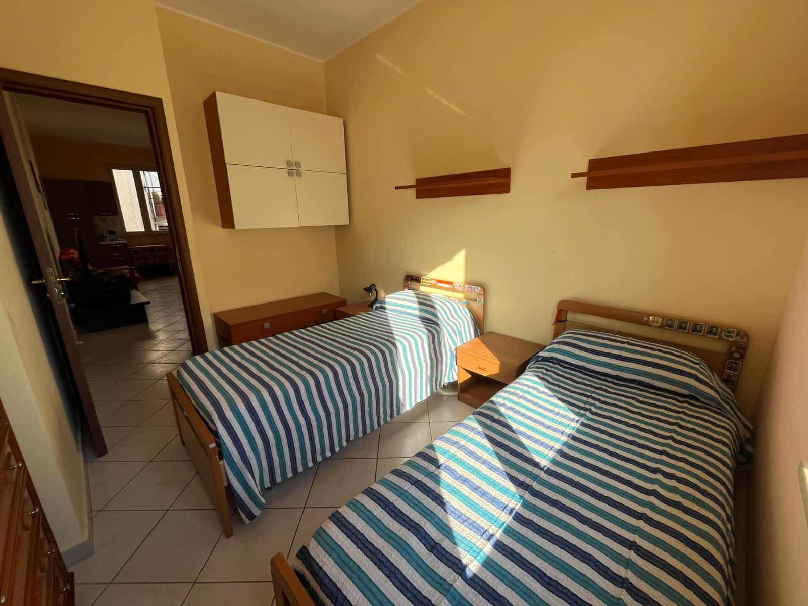 Letto singolo e letto matrimoniale in una camera da letto luminosa con pareti beige, armadio bianco e mensole in legno, ambiente accogliente e ben arredato, ideale per soggiorni confortevoli.