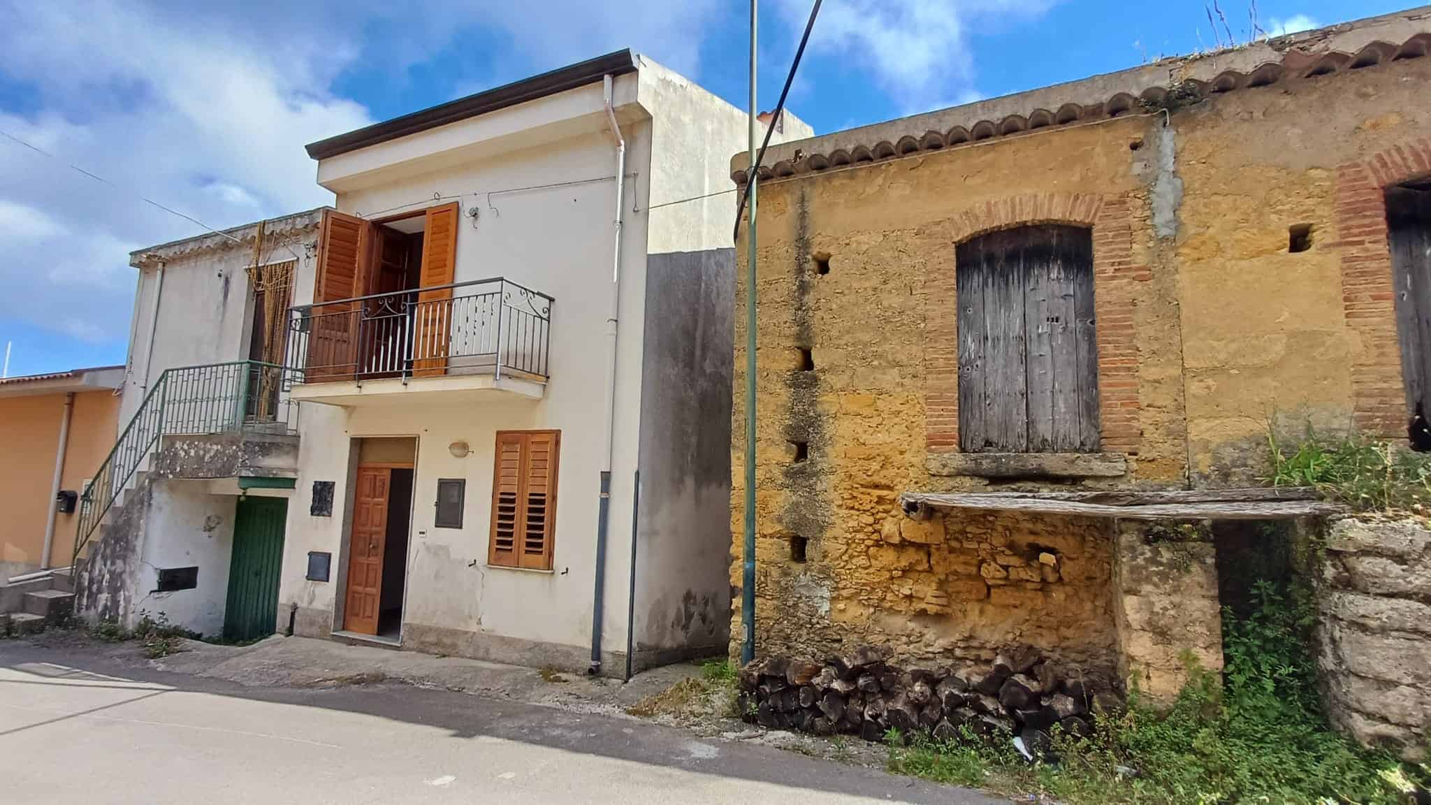 Antico edificio in pietra e moderna casa ristrutturata con balcone, situati in un quartiere storico di campagna, con cielo azzurro e nuvole bianche sullo sfondo.