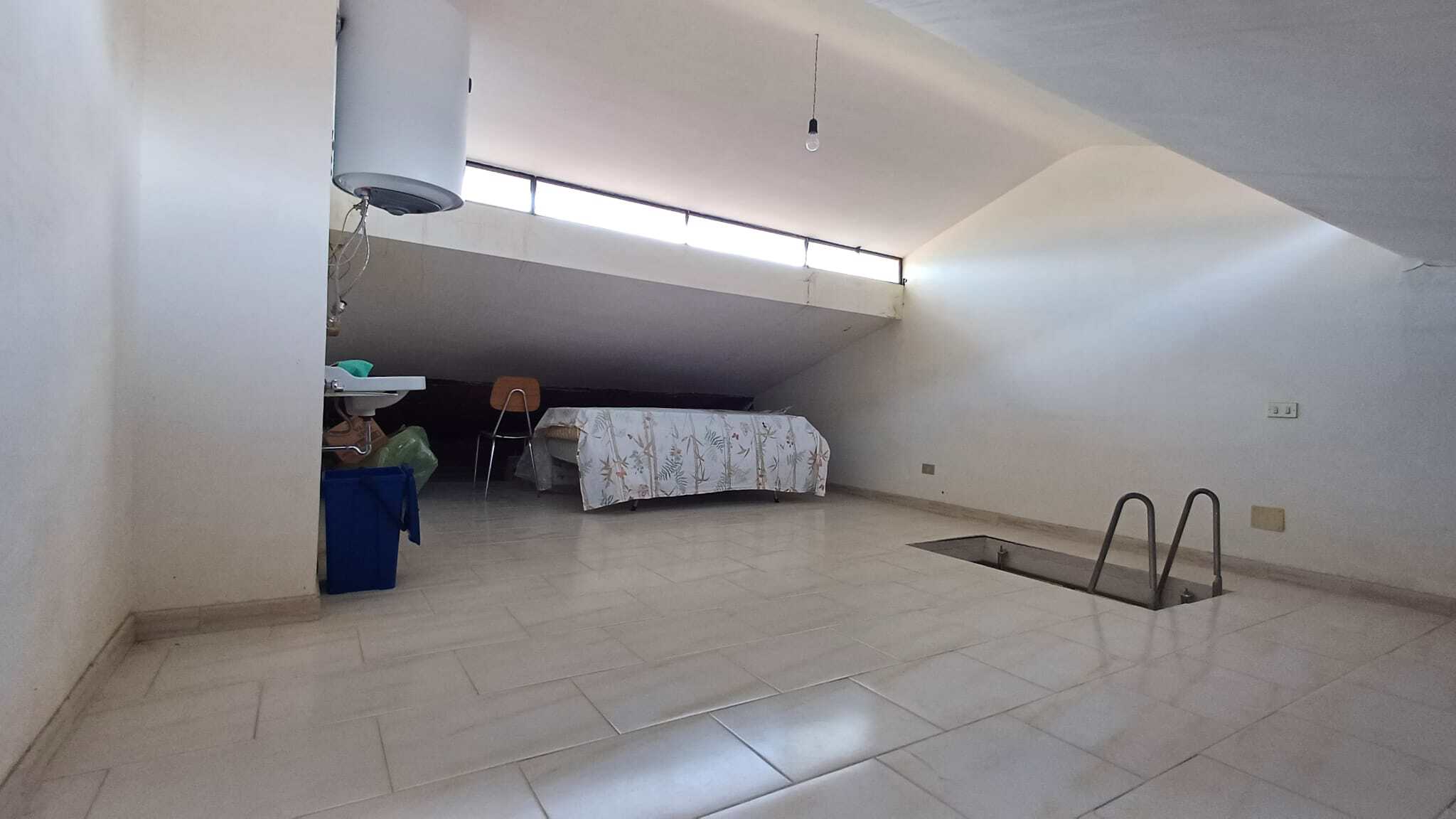 Ampio spazio in soffitta con pavimento in piastrelle chiare, area per riposo o studio, con letto e sedie, illuminata da luce naturale attraverso una finestra sul soffitto. Ideale per soluzioni di livi.
