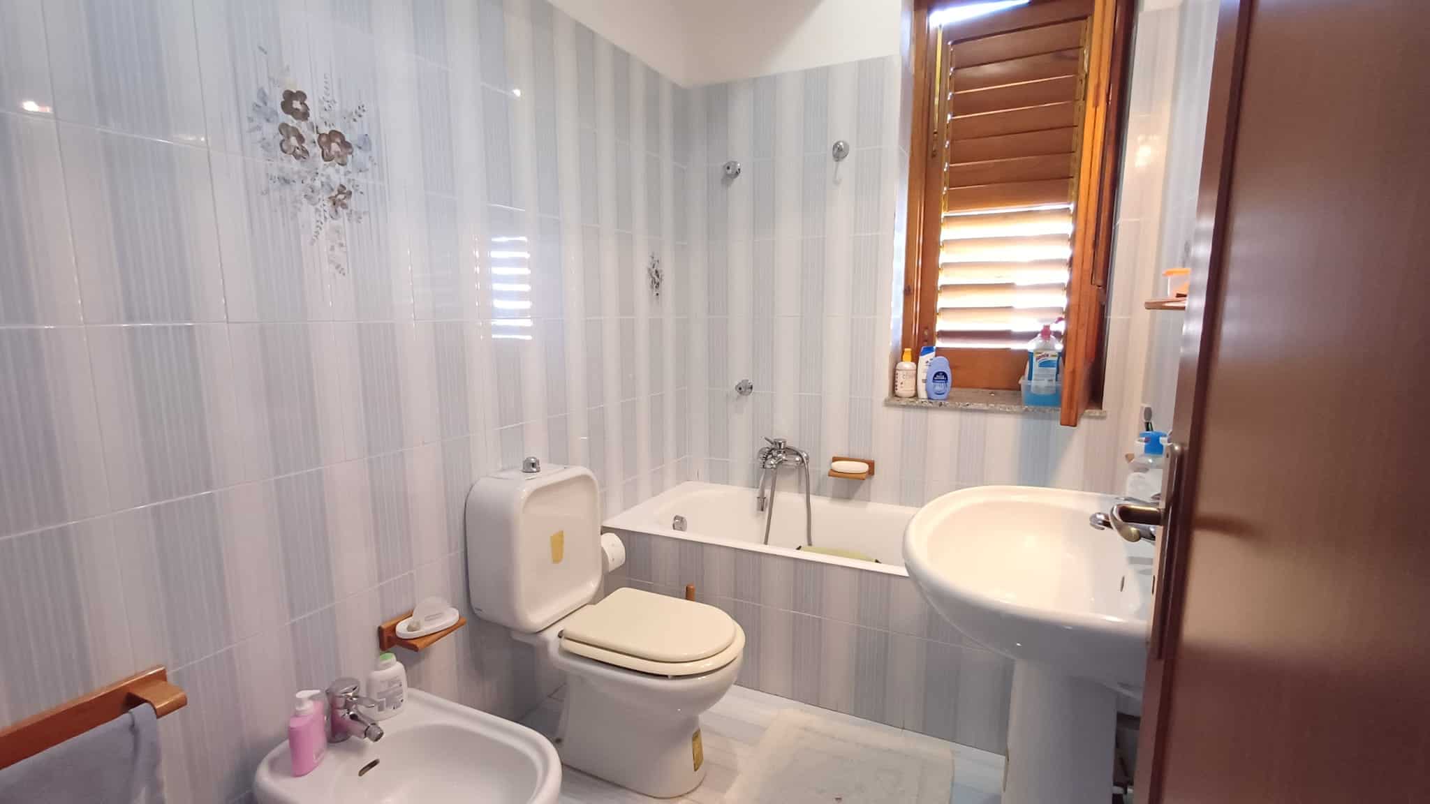 Il bagno di casa con vasca, lavabo e WC, illuminato da una finestra con persiane di legno, in stile semplice e funzionale, ideale per ambienti residenziali.