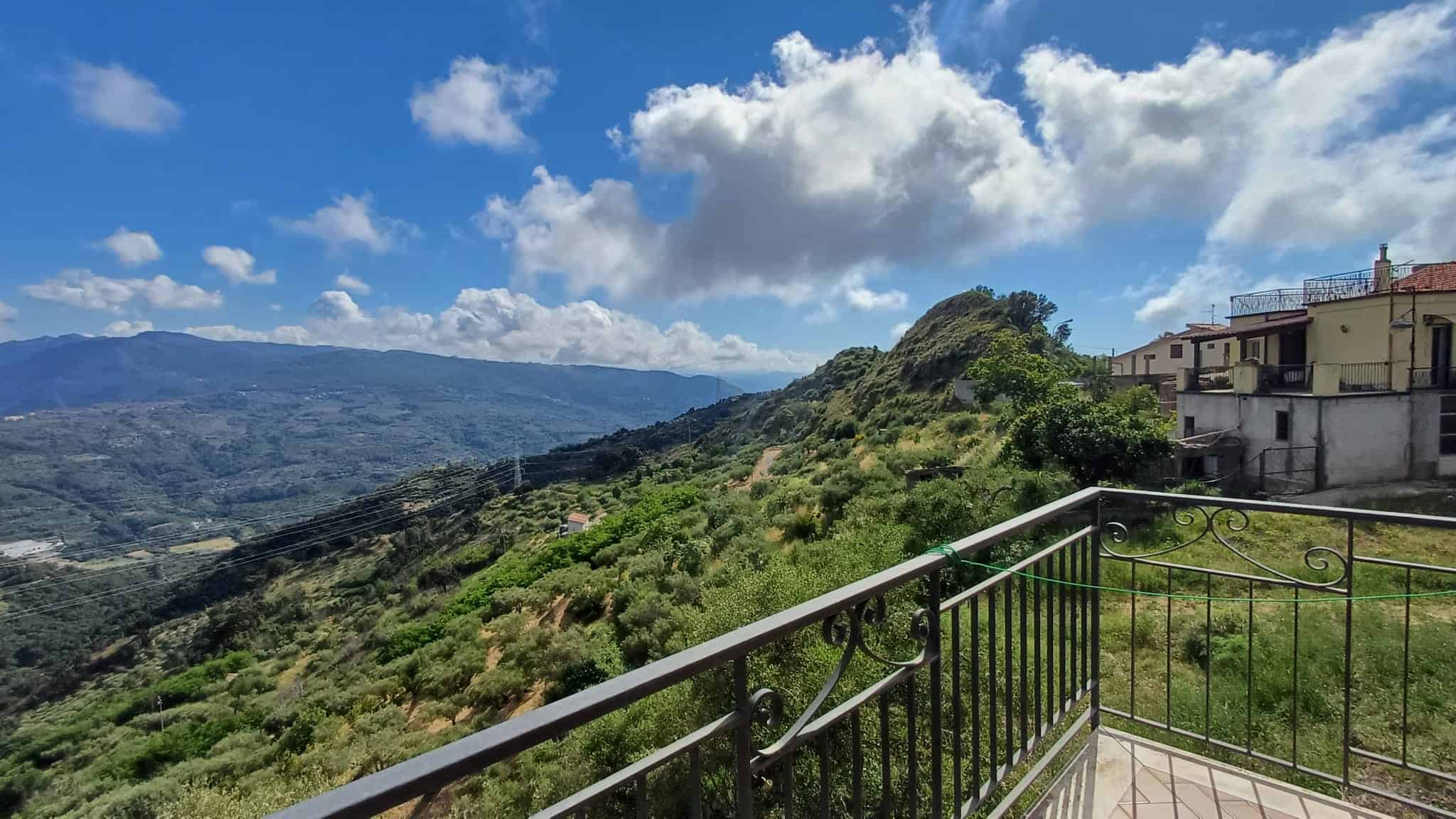 Godimento di una vista panoramica sulle colline verdi e il cielo nuvoloso dalla terrazza di una casa in collina, con un'area esterna in ferro battuto.