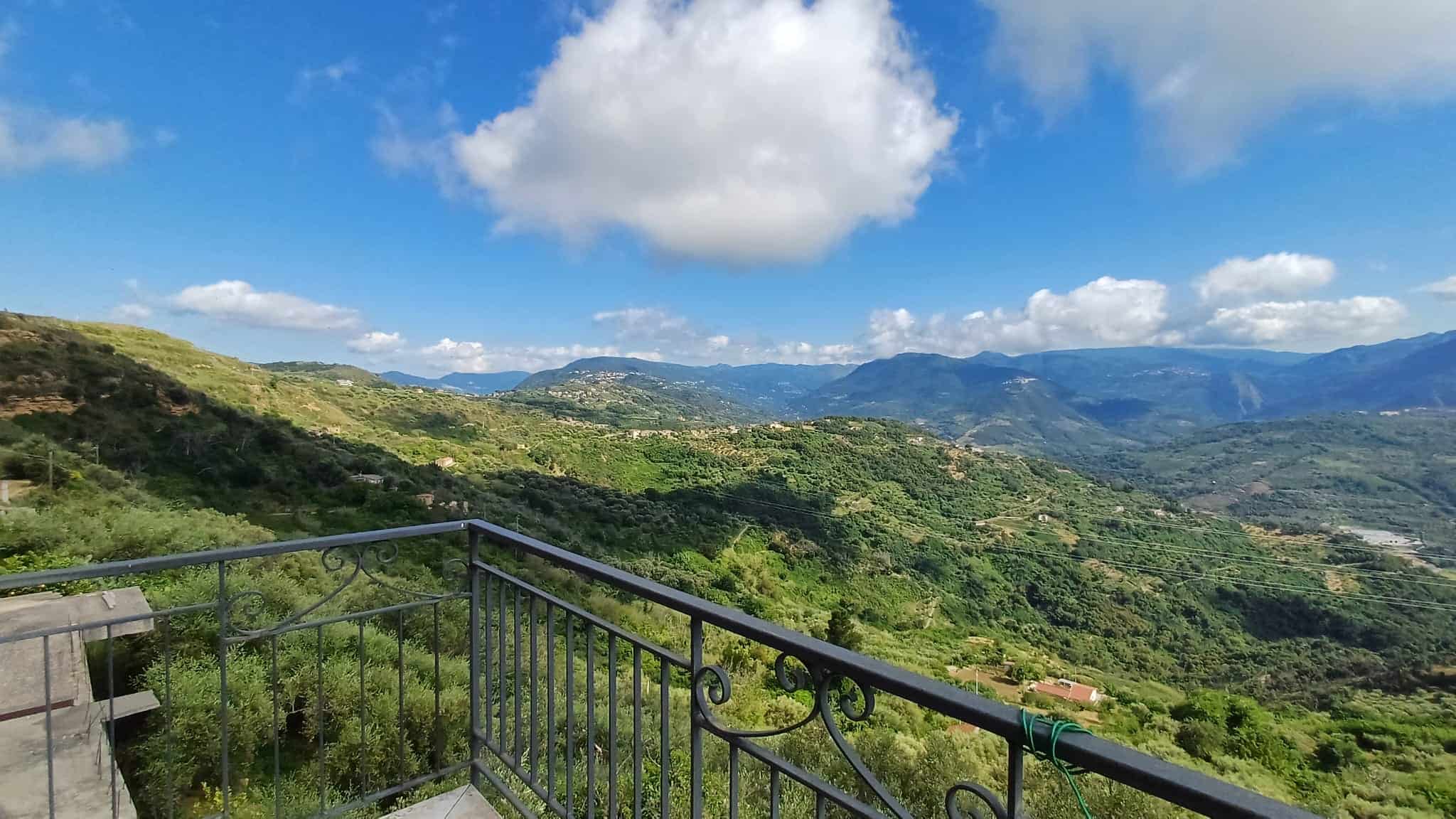 Vista panoramica delle colline verdi e delle montagne con un balcone in ferro battuto in primo piano, ideale per immobili di lusso e proprietà in vendita in zone panoramiche.