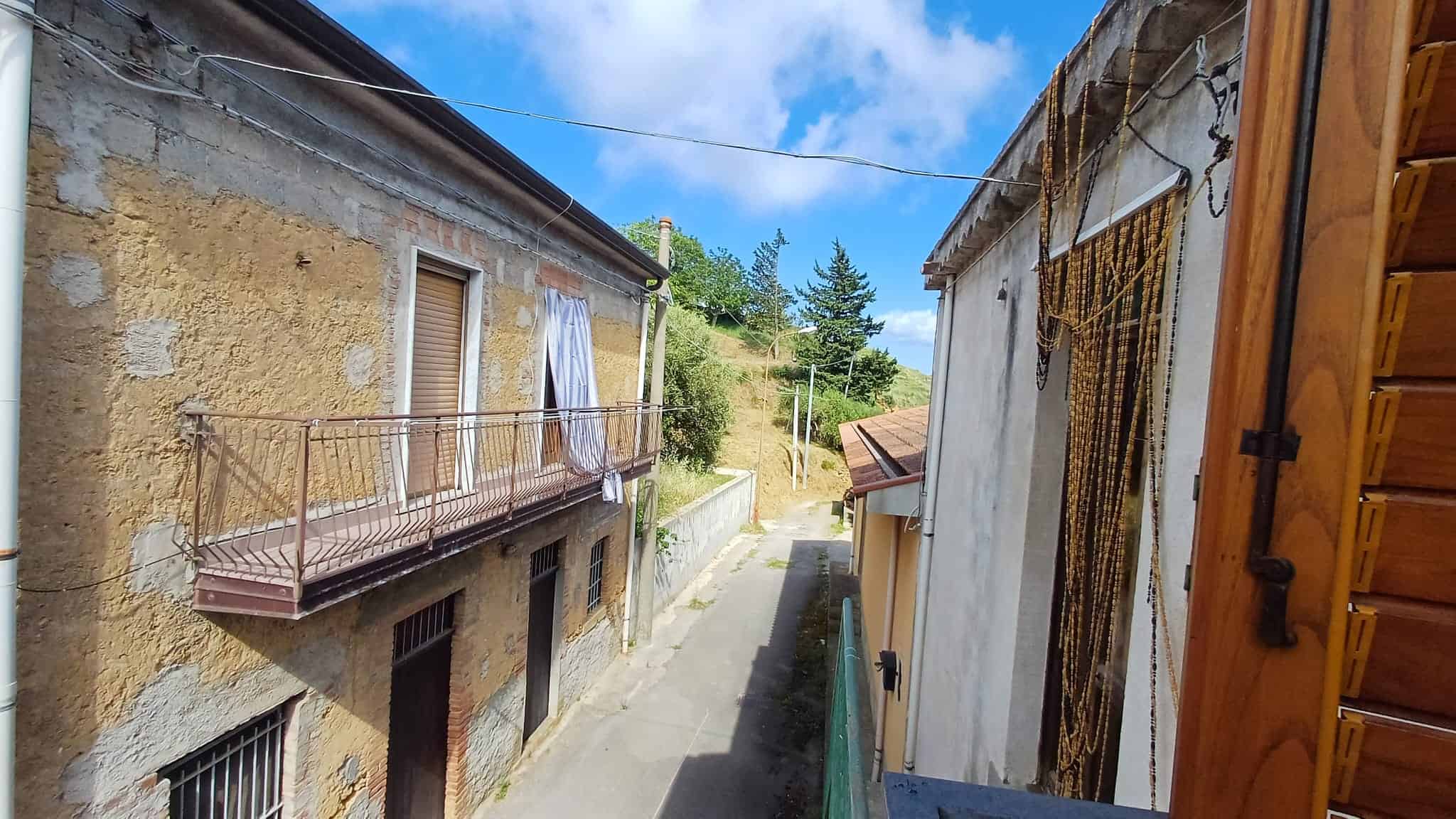 Scorcio di strada rurale tra case antiche in pietra e legno, con cielo azzurro e vegetazione verde, rappresentando un ambiente tranquillo e autentico in un quartiere storico italiano.