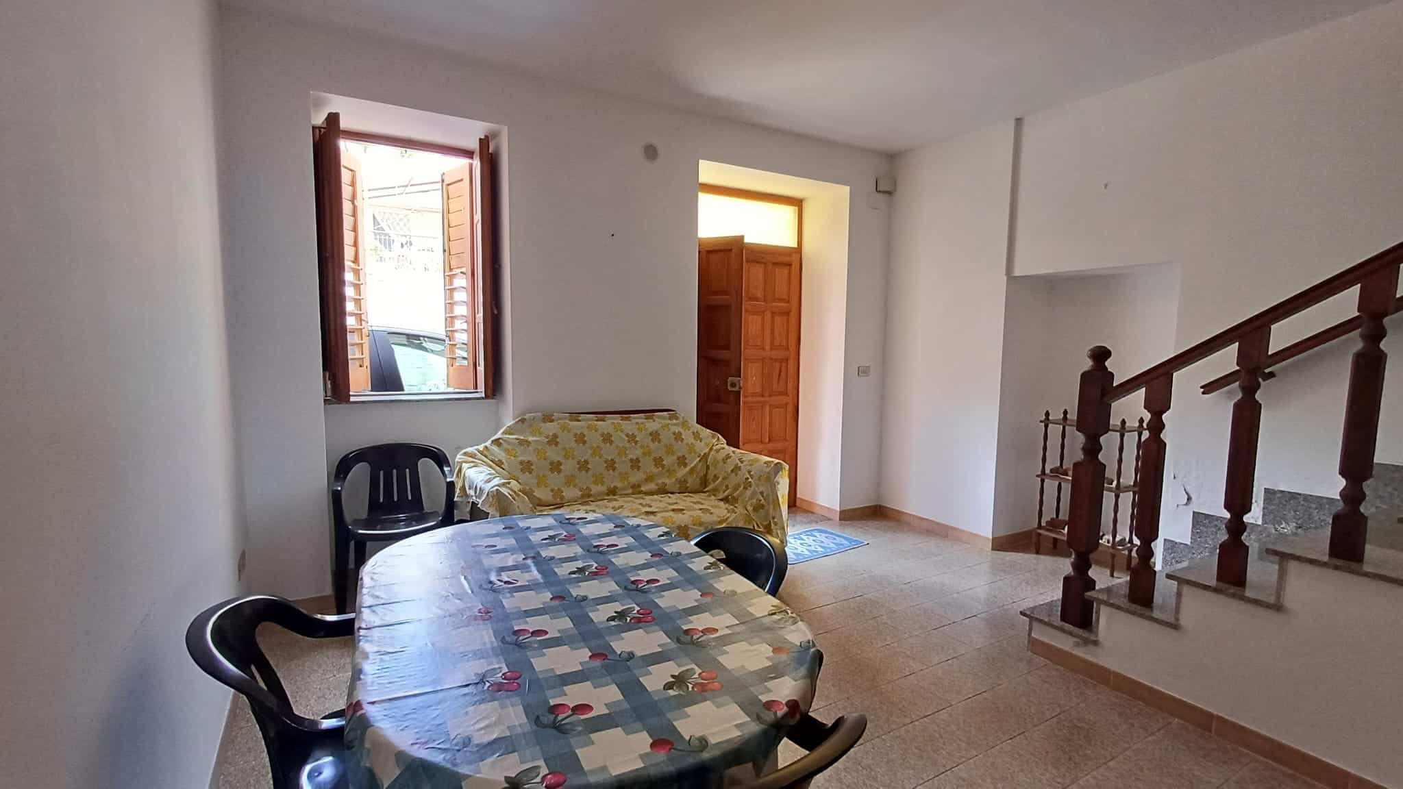 Interno di una casa con soggiorno luminoso, tavolo da pranzo rotondo, divano giallo e scale in legno, ambiente accogliente e spazioso, ideale per vivere e ricevere ospiti.