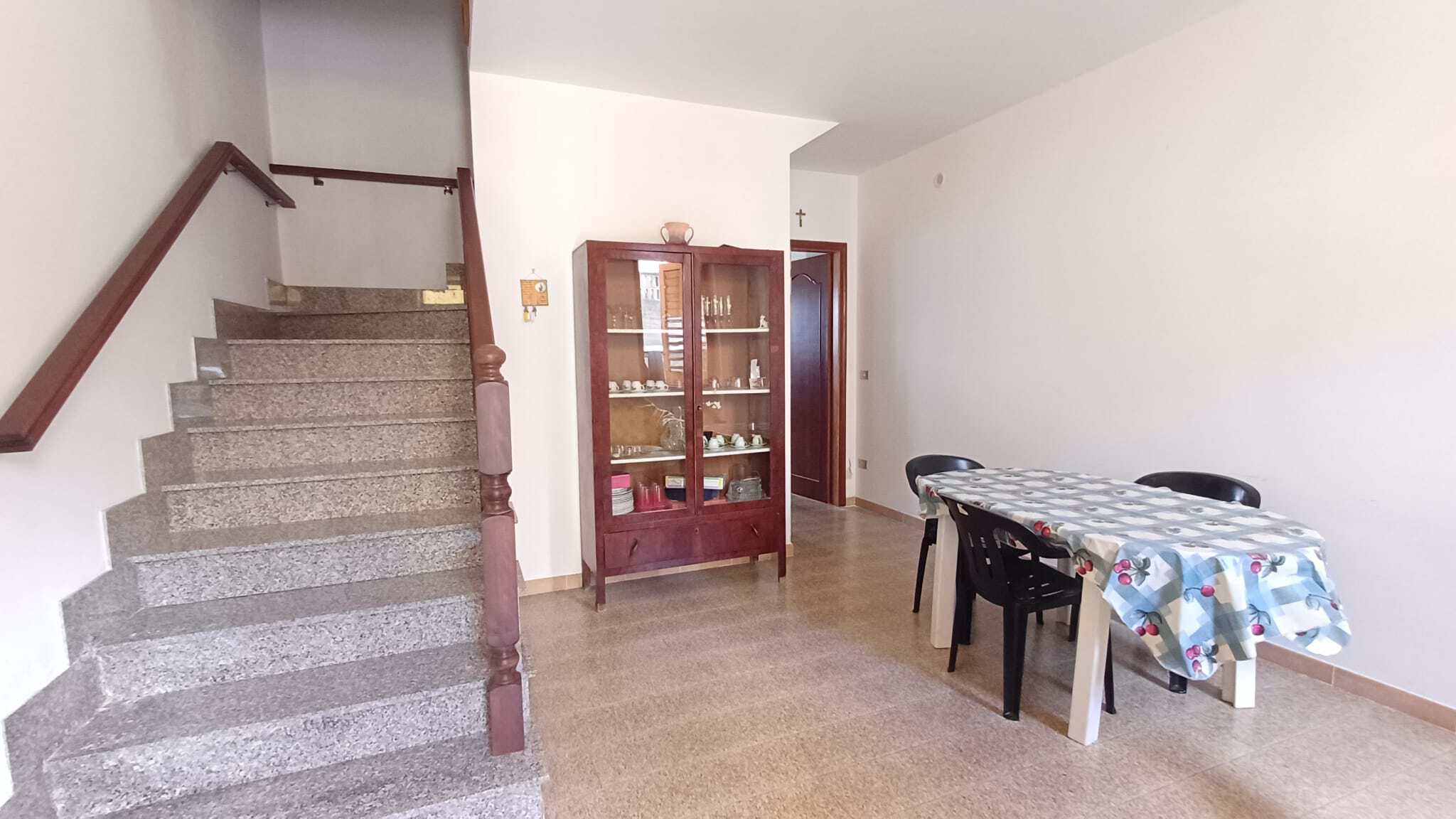 Ampia sala da pranzo con tavolo rettangolare e sedie nere, vicino a una parete bianca e a una vetrina in legno con oggetti decorativi, in un ambiente luminoso e accogliente.