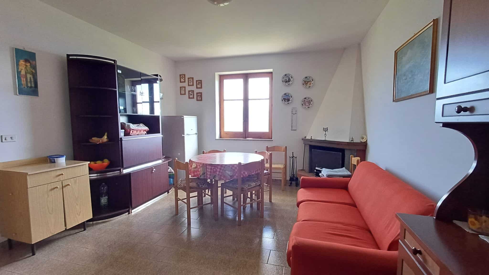 L'immagine mostra un soggiorno accogliente con mobili classici, un divano rosso, una zona pranzo con tavolo e sedie, e una finestra che lascia entrare luce naturale, creando un ambiente caldo e famili.