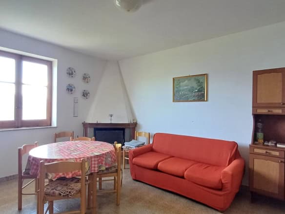 Ampia zona living con divano rosso, tavolo da pranzo rotondo con tovaglia colorata e finestra luminosa, arredamento semplice e accogliente, ideale per soggiorni confortevoli.