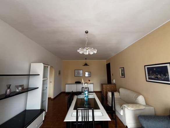 Ampia sala da pranzo con tavolo in vetro e sedie nere, soggiorno con divano bianco e pareti decorate con quadri, illuminazione centrale e arredi moderni, ambiente luminoso e accogliente.