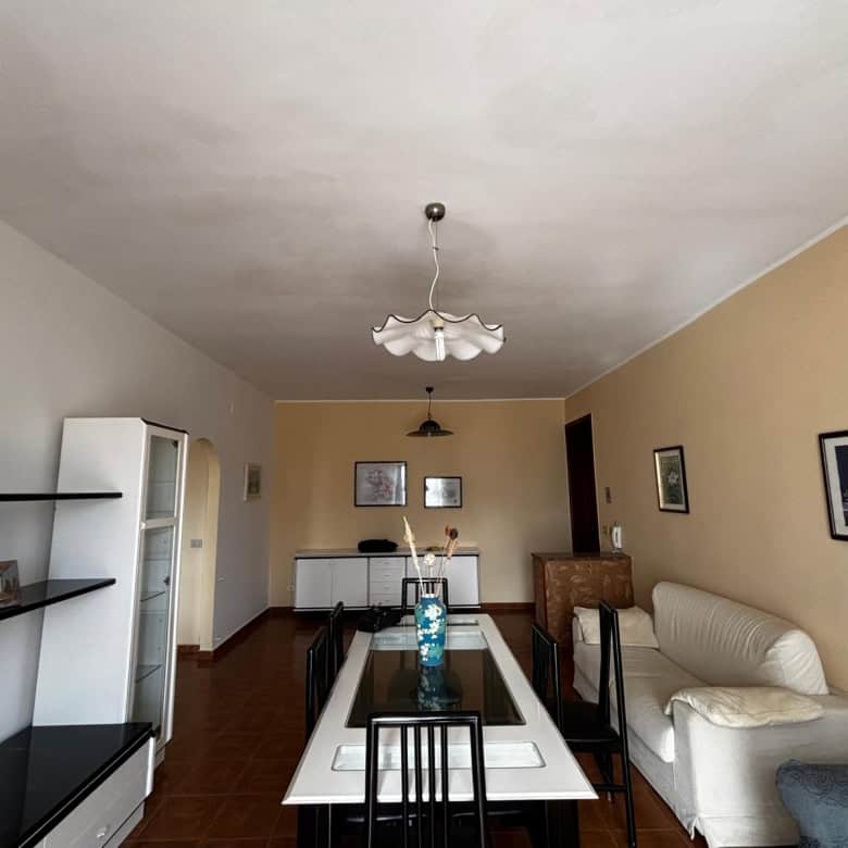Ampia sala da pranzo con tavolo in vetro e sedie nere, soggiorno con divano bianco e pareti decorate con quadri, illuminazione centrale e arredi moderni, ambiente luminoso e accogliente.