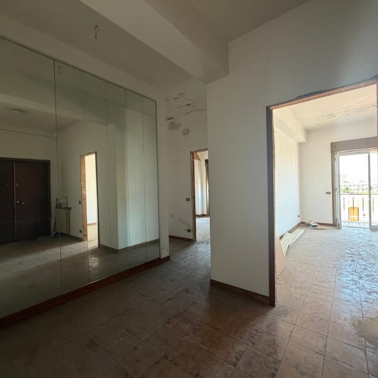 Interno di un appartamento in fase di ristrutturazione con pareti bianche, grandi finestre e pavimento in piastrelle, pronto per essere arredato.