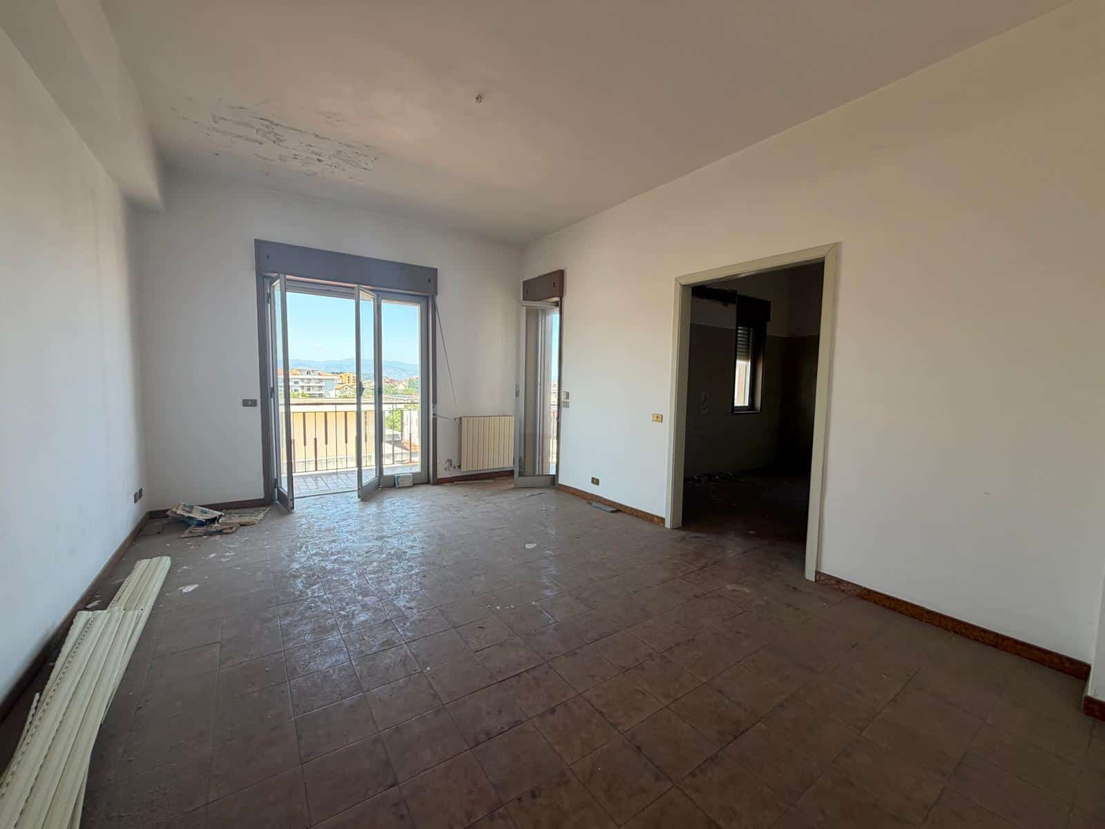 Ampio soggiorno vuoto con grandi finestre e balcone, luce naturale, pavimento in piastrelle, pareti bianche, vista panoramica sulla città, ideale per ristrutturazione e arredamento moderno.