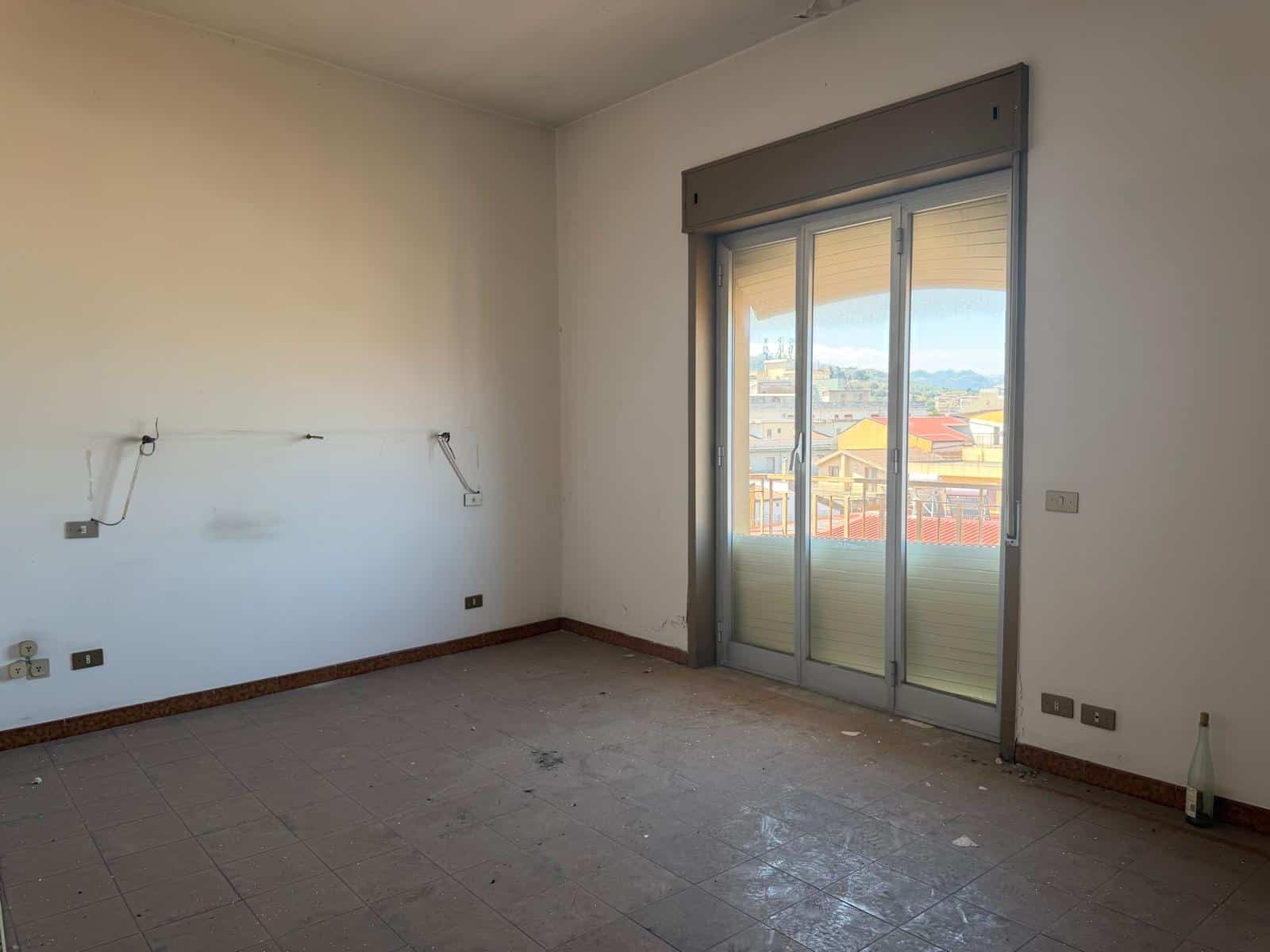Stanza vuota in fase di ristrutturazione con porta finestra che dà su un balcone e vista panoramica sulla città.