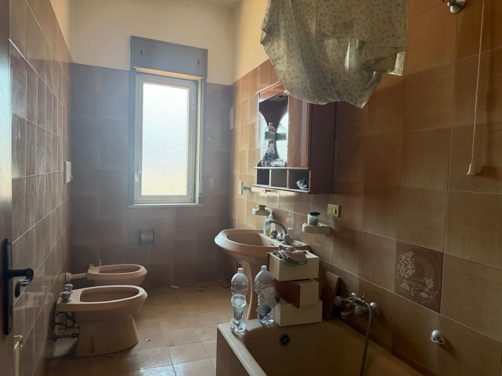 Il bagno con bidet e lavabo, finestra luminosa e pareti rivestite in piastrelle marroni, con alcuni oggetti e bottiglie d'acqua sul mobile, in un ambiente domestico semplice e funzionale.