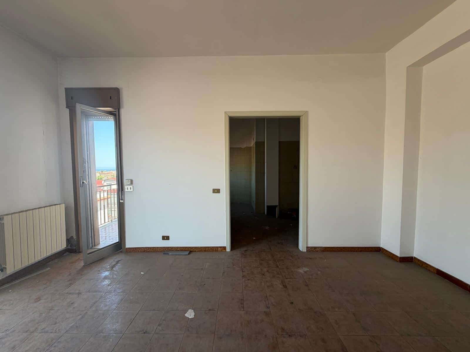 Interno di un appartamento vuoto con pareti bianche, porta finestra e balcone, pronto per essere arredato, in una zona residenziale soleggiata.