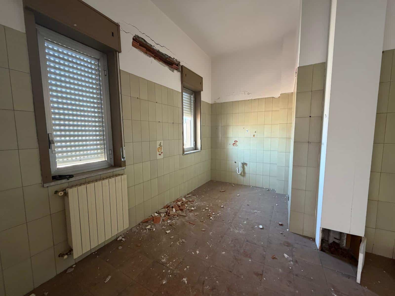 Una stanza vuota in fase di ristrutturazione con pareti rivestite di piastrelle beige, due finestre con tapparelle, un radiatore e pavimento sporco con detriti.