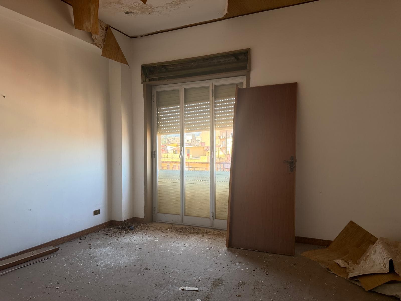 Stanza vuota in fase di ristrutturazione con porta e finestra, pavimento in cemento, luce naturale, pronto per arredamento e personalizzazione.
