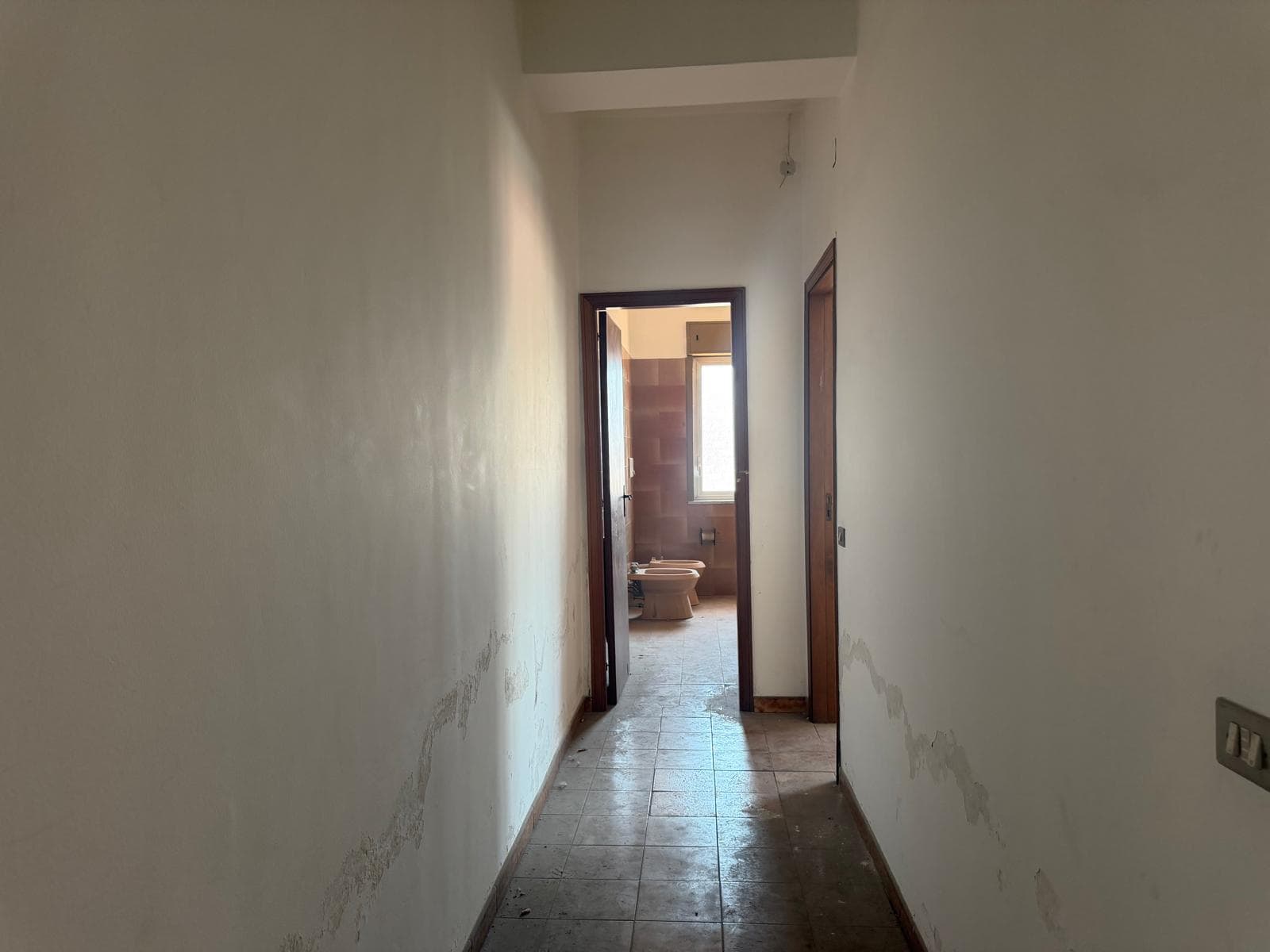 Corridoio interno di un appartamento in fase di ristrutturazione con pareti danneggiate e pavimento in piastrelle, pronto per lavori di rinnovamento e decorazione.