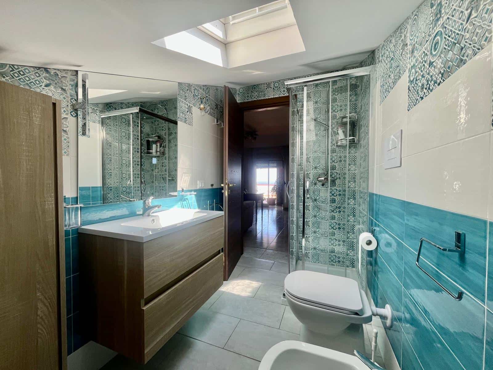 Lavabo moderno con specchio e doccia separata in bagno luminoso, arredato con stile minimalista e piastrelle decorative in tonalità di blu e bianco, ideale per ambienti di comfort e funzionalità.