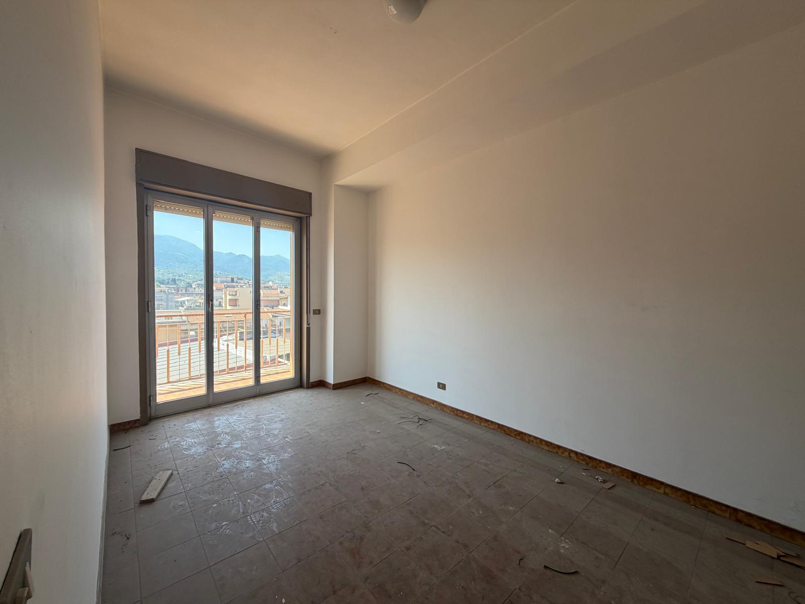 Ampia stanza vuota con pareti bianche e grande finestra scorrevole che offre vista sulla città e sulle montagne circostanti, pronta per essere arredata e personalizzata.