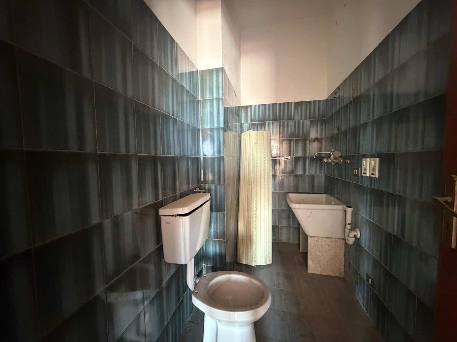 Lavabo e servizi igienici in un bagno con pareti rivestite di piastrelle blu scuro, con accessori essenziali e un ambiente funzionale.
