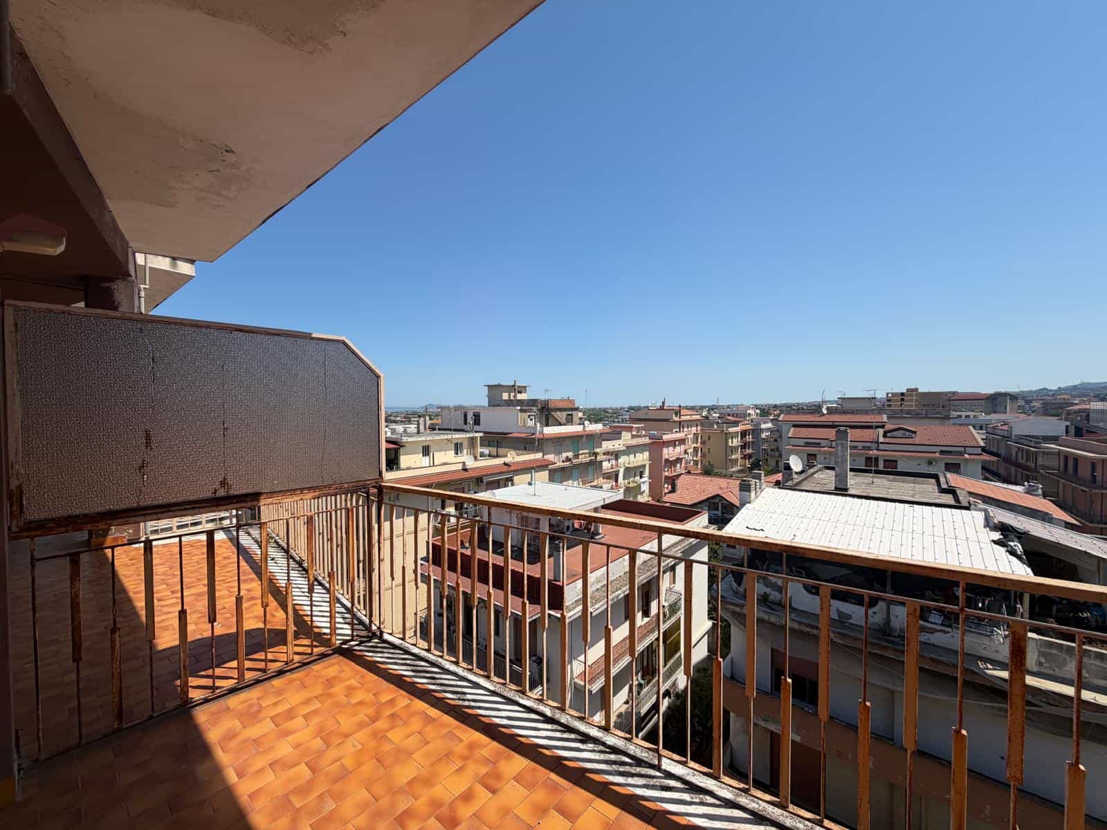 Balcone di un appartamento con vista sulla città, con ringhiera in metallo e pavimento in piastrelle arancioni, sotto un cielo sereno e azzurro, in un contesto urbano residenziale.