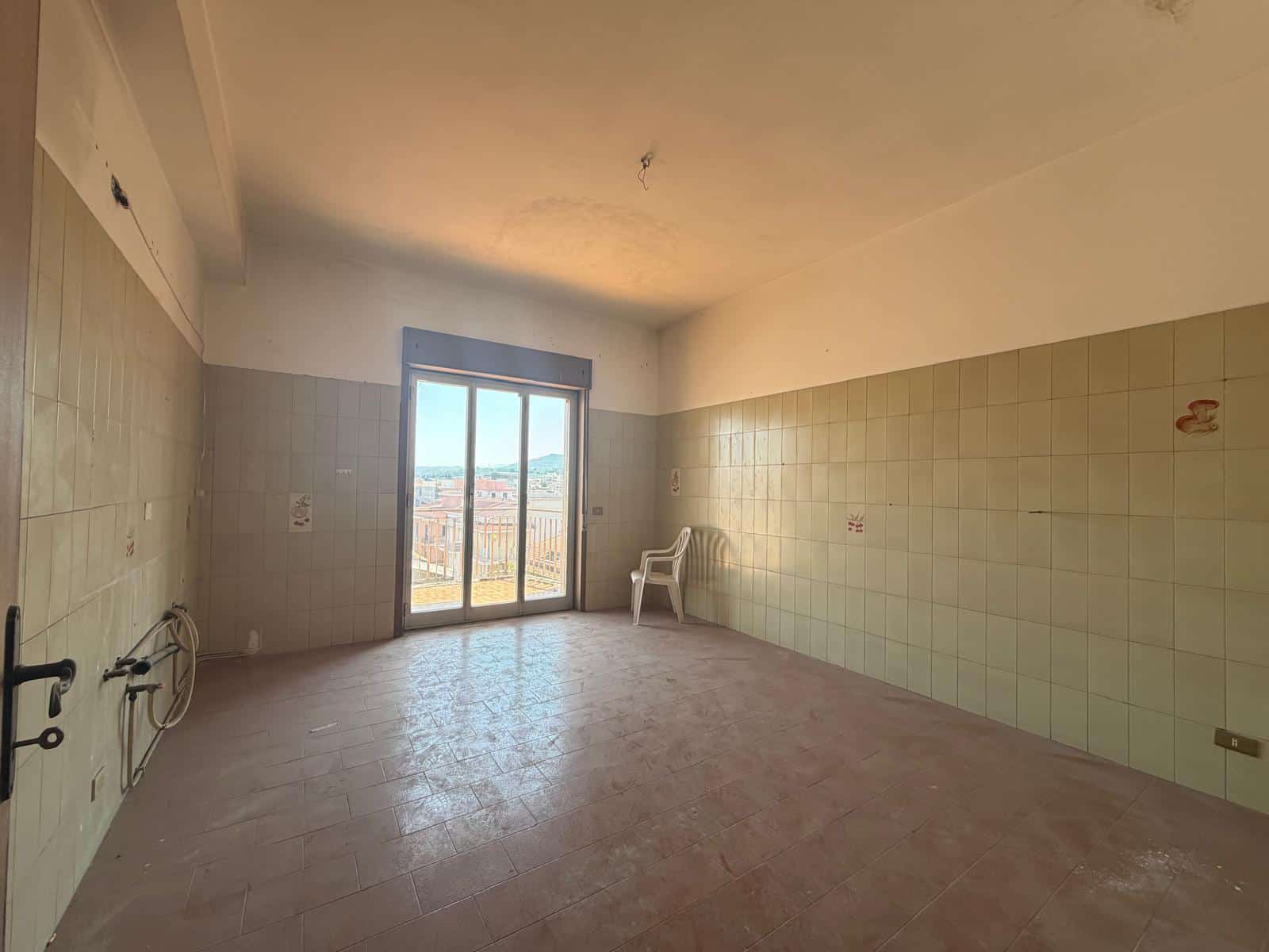 Una stanza vuota con pareti rivestite di piastrelle beige, grande finestra con balcone e vista sulla città, pavimento in cemento e una sedia bianca, pronta per essere ristrutturata o personalizzata.