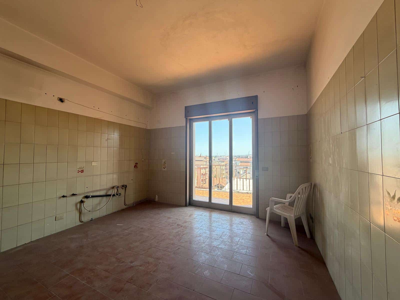 Stanza vuota con pareti rivestite di piastrelle, grande finestra con vista sulla città, pavimento in piastrelle e sedia bianca, ambiente luminoso e spazioso.