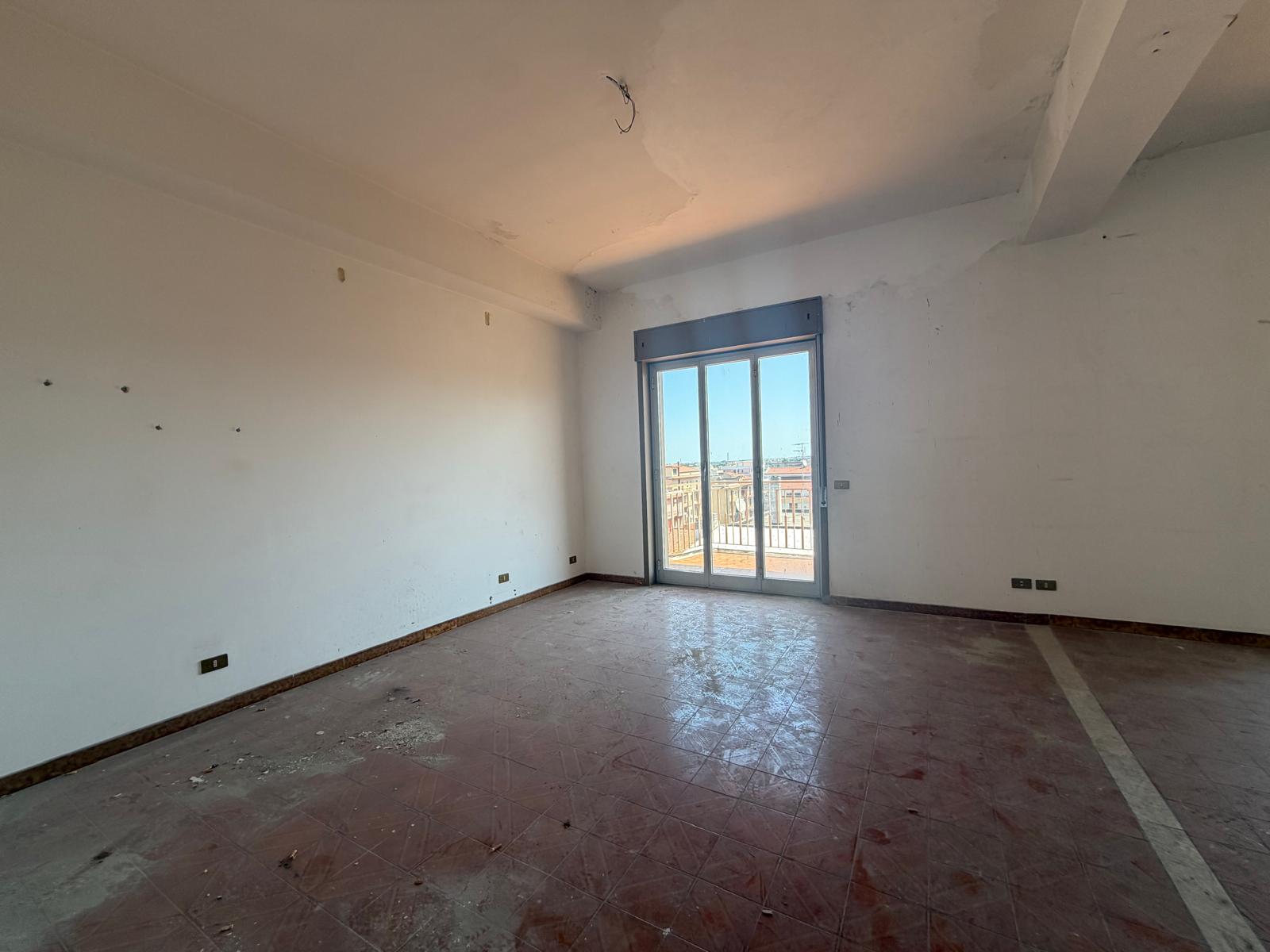 Interno di un appartamento in fase di ristrutturazione con pareti bianche, pavimento in ceramica e grande finestra che permette l'ingresso di luce naturale.