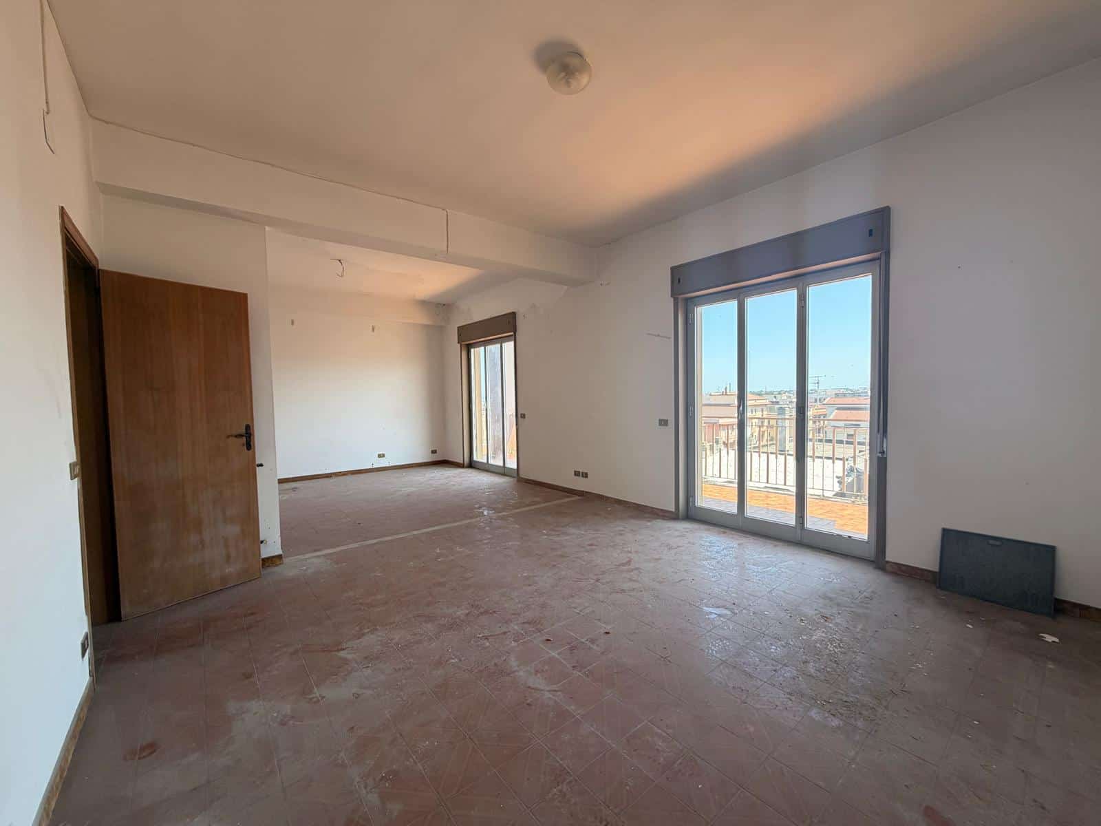 Interno di un appartamento vuoto con grandi finestre e vista sulla città, pronto per essere arredato, in fase di ristrutturazione, con pareti bianche e pavimento in cemento.