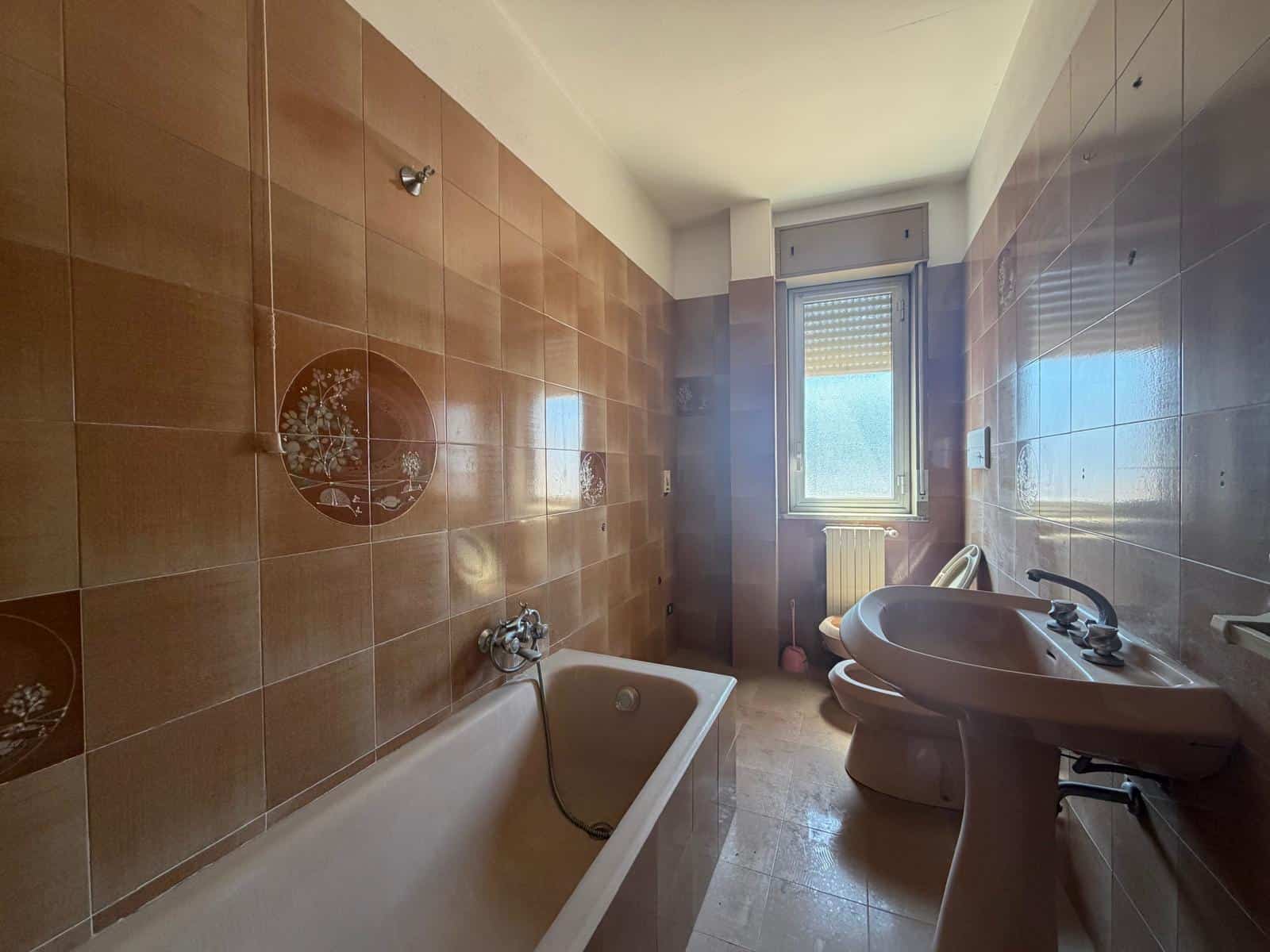 Bagno con vasca e lavabo in un appartamento, illuminato dalla luce naturale proveniente dalla finestra. Pareti rivestite di piastrelle marroni e pavimento in ceramica chiara, ambiente pulito e funzion.