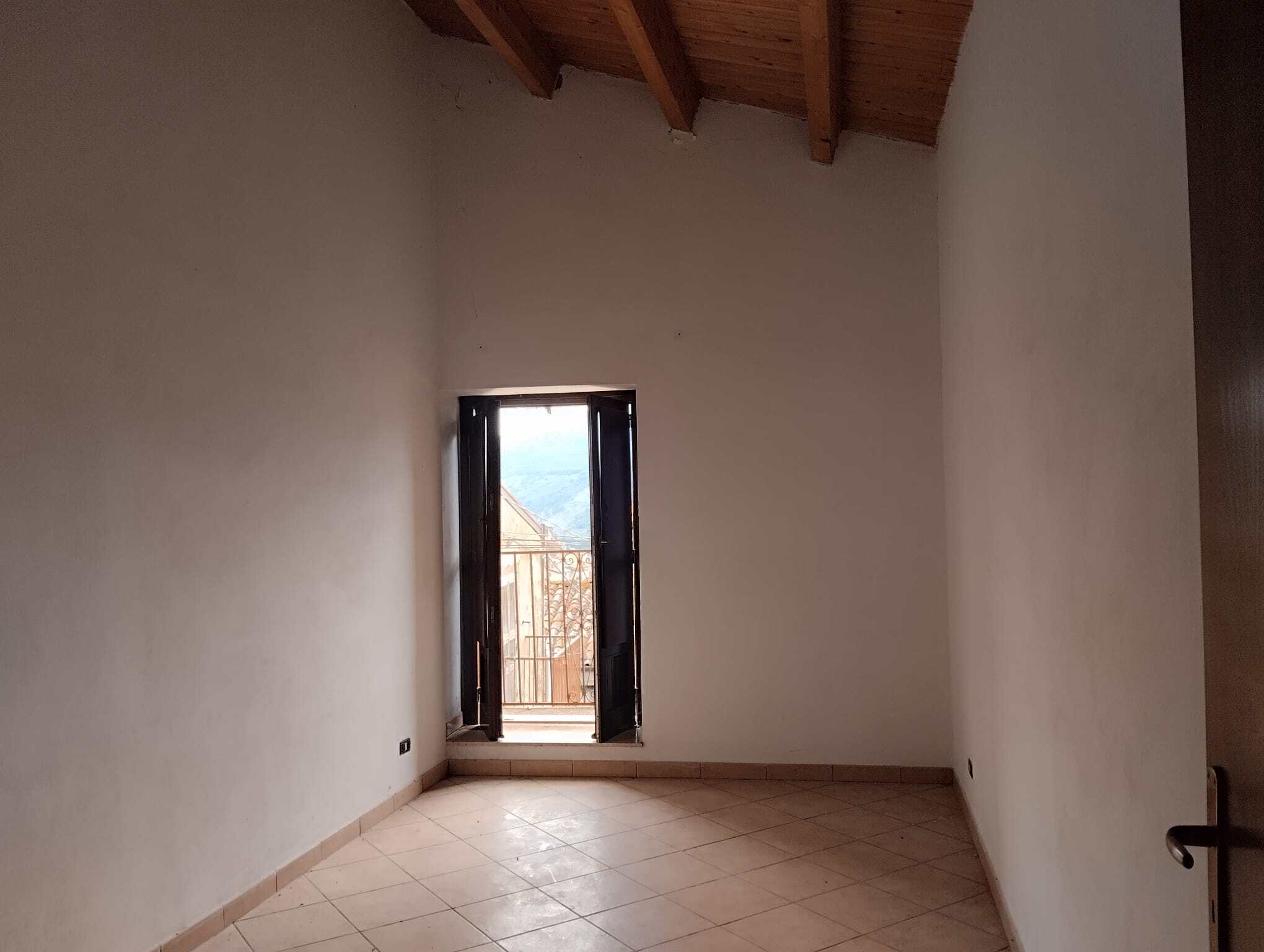 Ampia stanza vuota con pareti bianche, pavimento in piastrelle e porta finestra che conduce a un balcone con vista sulle montagne, pronta per essere arredata e personalizzata.