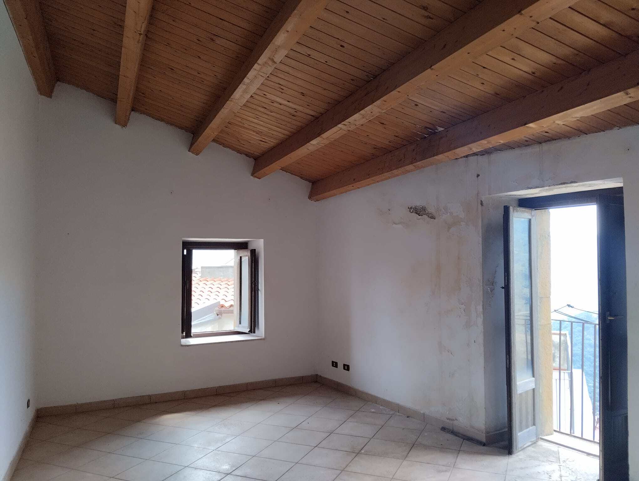 Interno di un appartamento in fase di ristrutturazione con soffitto in legno e pareti bianche, finestra e porta balcone che affacciano su un panorama urbano.