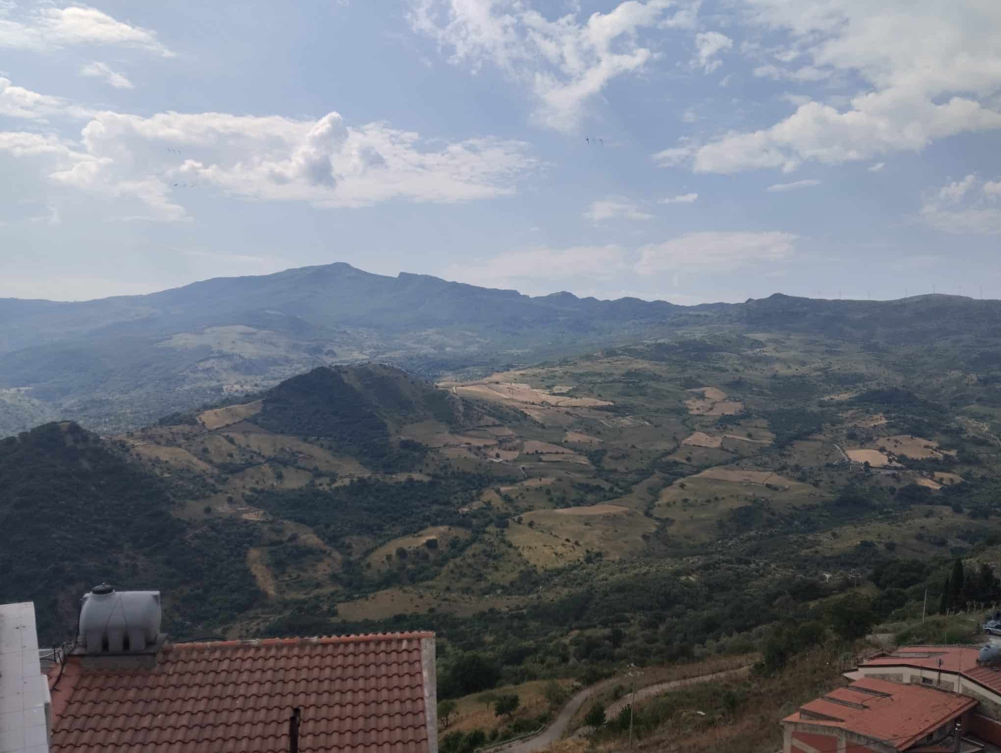 VISTA PANORAMICA DI UN PAESAGGIO MONTANO CON COLLINE VERDI E CIELO PARZIALMENTE NUVOLOSO, vista da una zona residenziale con tetti rossi in primo piano, ideale per immobili e terreni in vendita in zon.