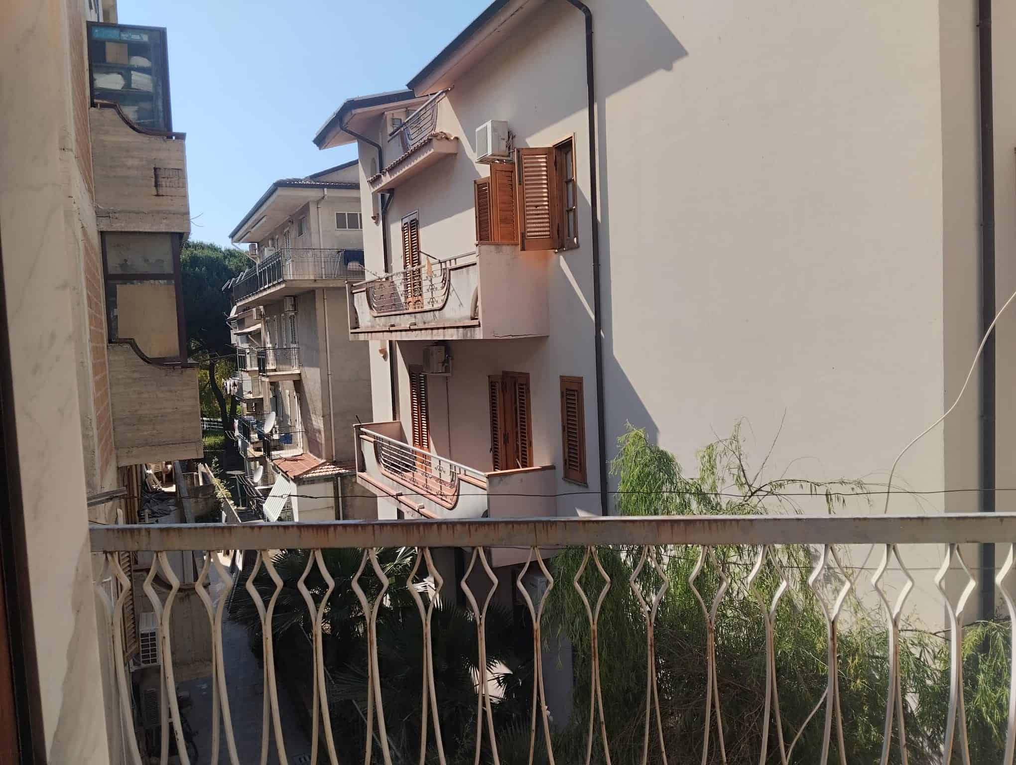 Vicina di case residenziali con balcone in ferro battuto, vista su strada e edifici circostanti, illuminata dalla luce solare, in un quartiere tranquillo e ben curato.