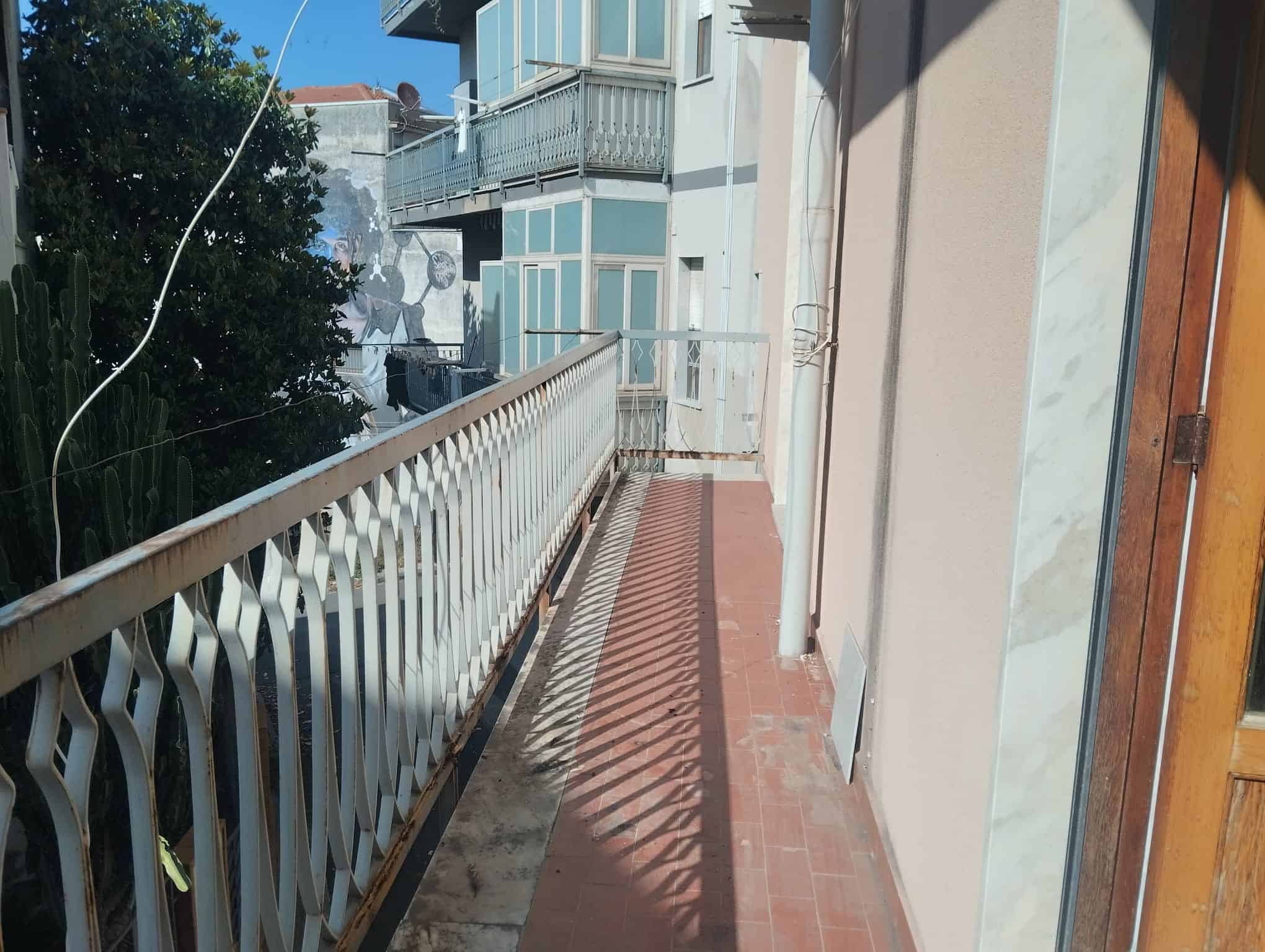 Finestra e balcone di un appartamento con ringhiera in ferro battuto, vista su edifici residenziali e cielo sereno, in un contesto urbano italiano.