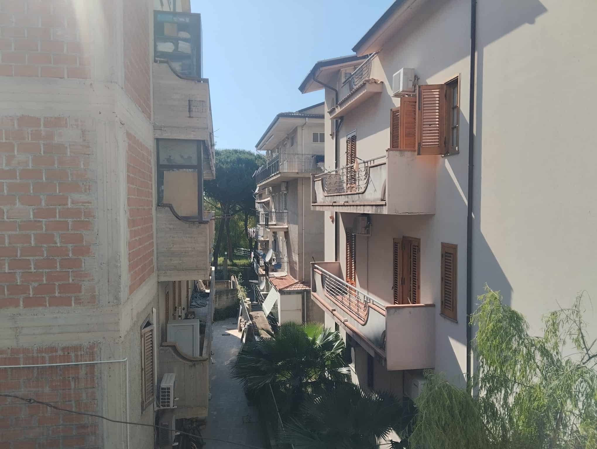 Vista di un vicolo residenziale con edifici multifamiliari, balconi e piante verdi, in un quartiere tranquillo e soleggiato di città italiana.