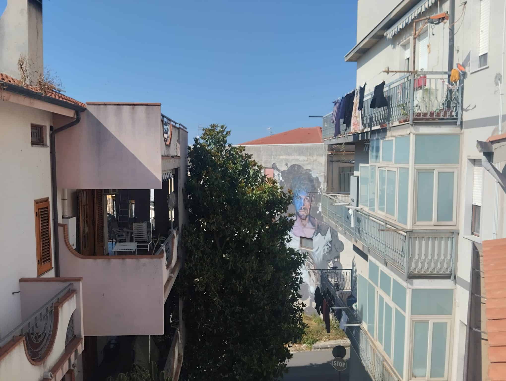 Vista urbana di un vicolo residenziale con edifici moderni e un grande albero centrale, in una giornata soleggiata, in Italia.
