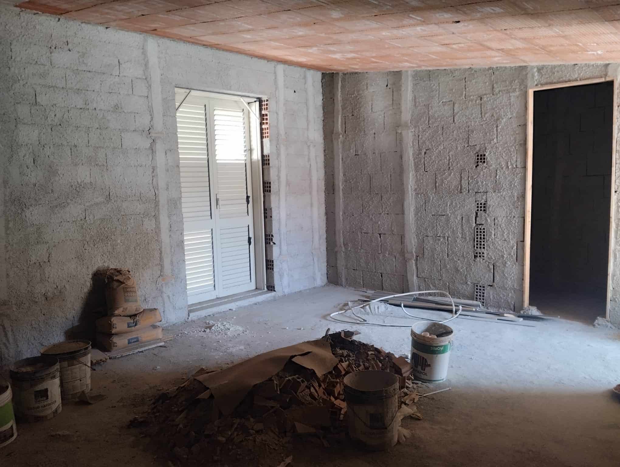 Interno di un appartamento in costruzione con pareti in cemento, pavimento in fase di finitura e materiali da costruzione visibili.