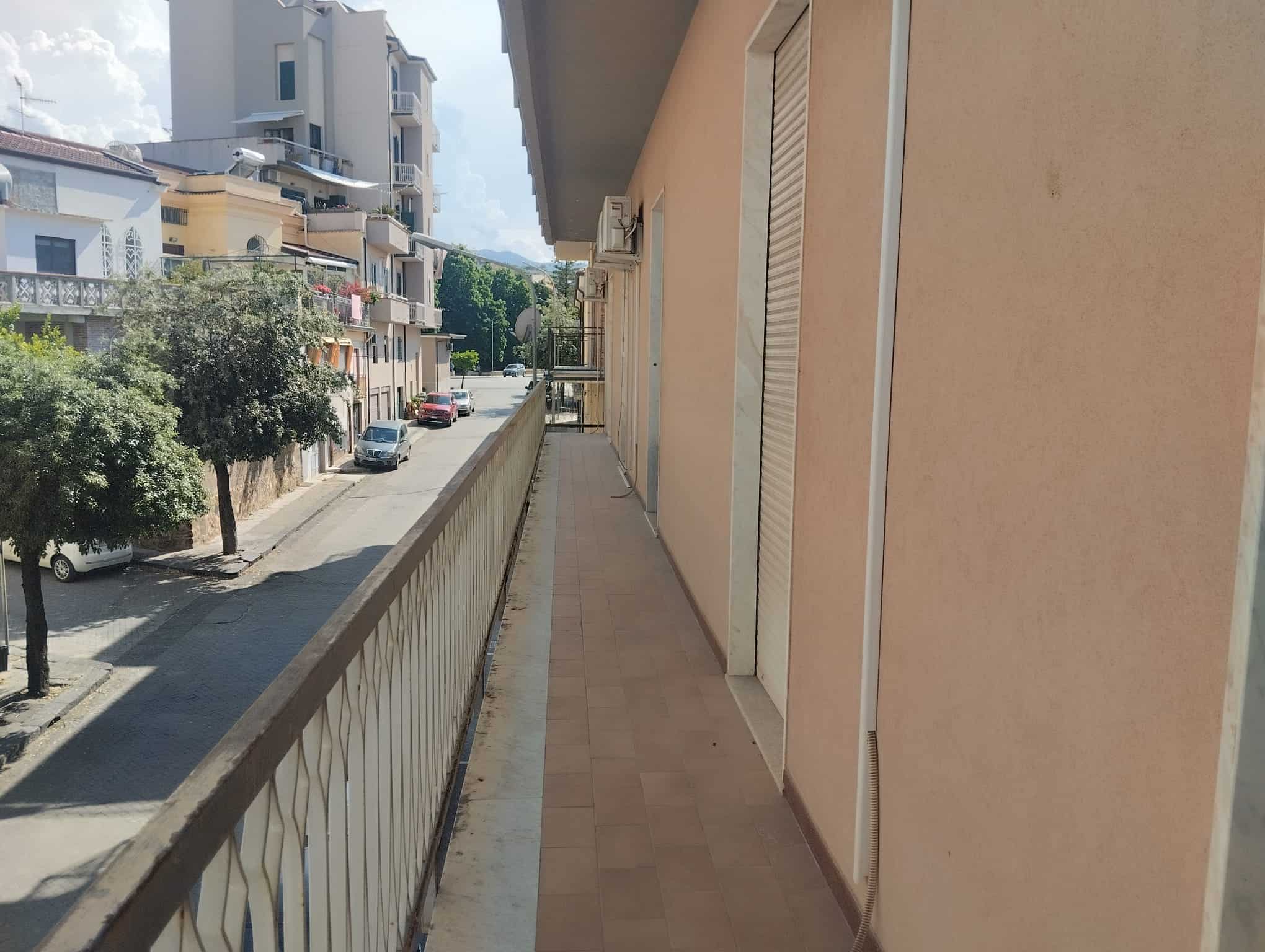 Balcone di un appartamento con vista sulla strada e sugli edifici circostanti, in un contesto urbano residenziale, con ringhiera in ferro e pavimentazione in piastrelle beige, in una giornata soleggia.
