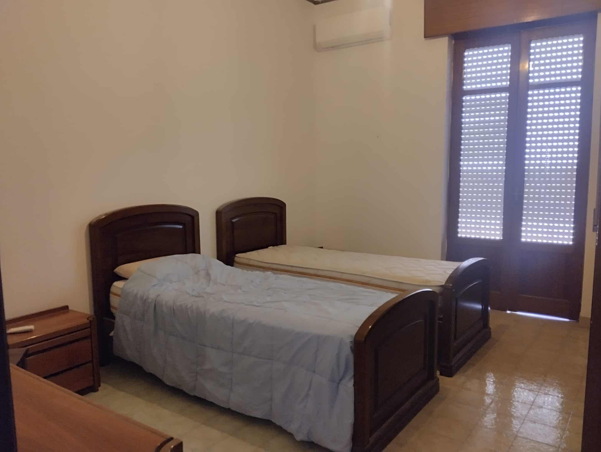 Camera da letto con due letti singoli in stile classico, finestra con persiane in legno, arredamento semplice e funzionale, ambiente luminoso e accogliente.