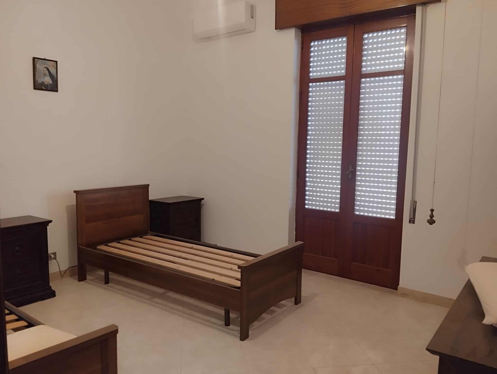 Il letto singolo in legno in una camera da letto luminosa con porta finestra in legno e persiane, arredamento semplice e funzionale, ideale per affitti brevi o uso domestico.