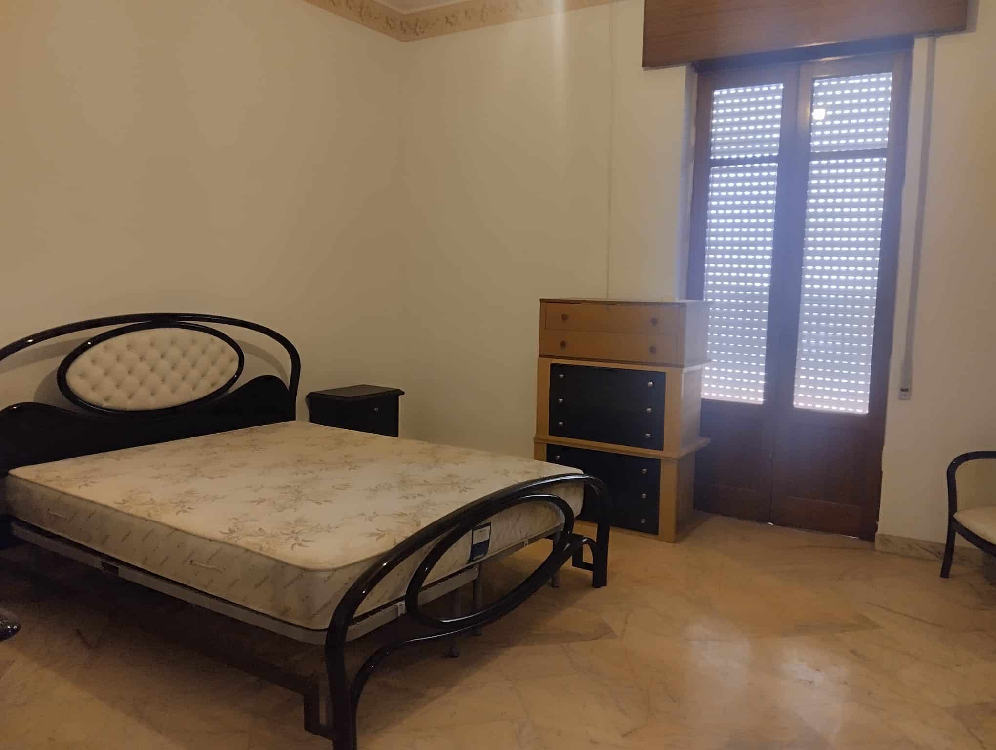 Un letto matrimoniale in una camera da letto semplice con pareti beige, una finestra con persiane e mobili in legno, ambiente accogliente e funzionale.