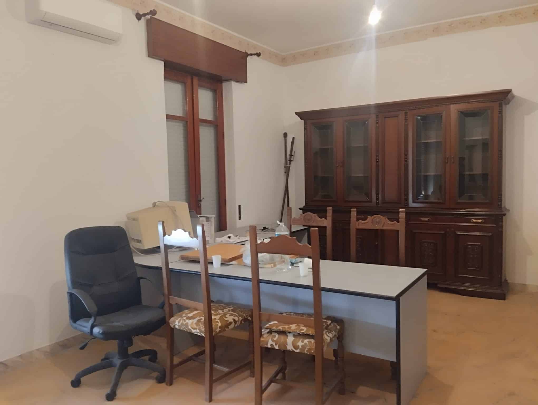 Un'area ufficio con scrivania, sedia ergonomica e sedie in legno, con una grande credenza in legno scuro e una finestra con tende marroni, in un ambiente domestico ben illuminato.