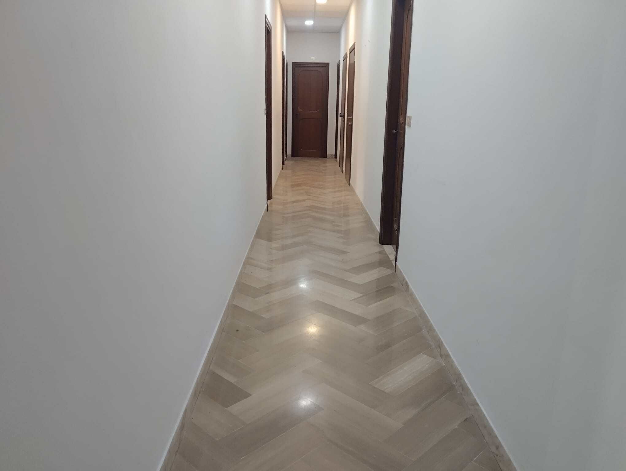 Aggiungi un'immagine di un corridoio interno di una casa con pareti bianche e pavimento in legno chiaro, con porte in legno scuro ai lati e illuminazione soffusa.