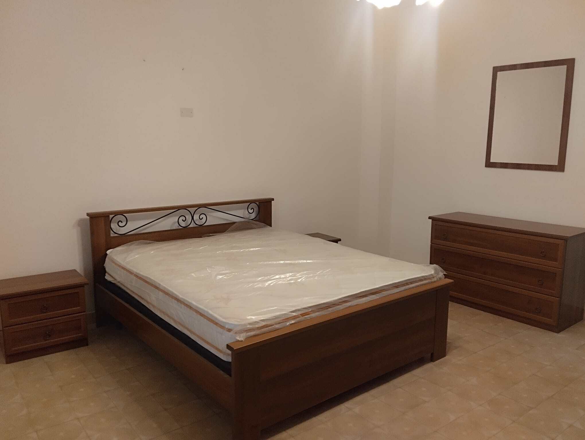 Un letto matrimoniale in legno con due comodini e una cassettiera in una stanza luminosa e minimalista, ideale per affitti a breve termine o residenze permanenti, con pareti bianche e pavimento in pia.