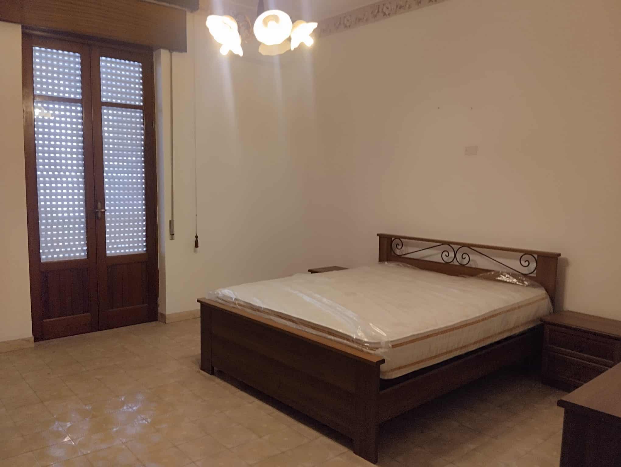 Ampia camera da letto con letto matrimoniale in legno, finestra con persiane e illuminazione soffusa, ideale per affitti turistici o residenziali.