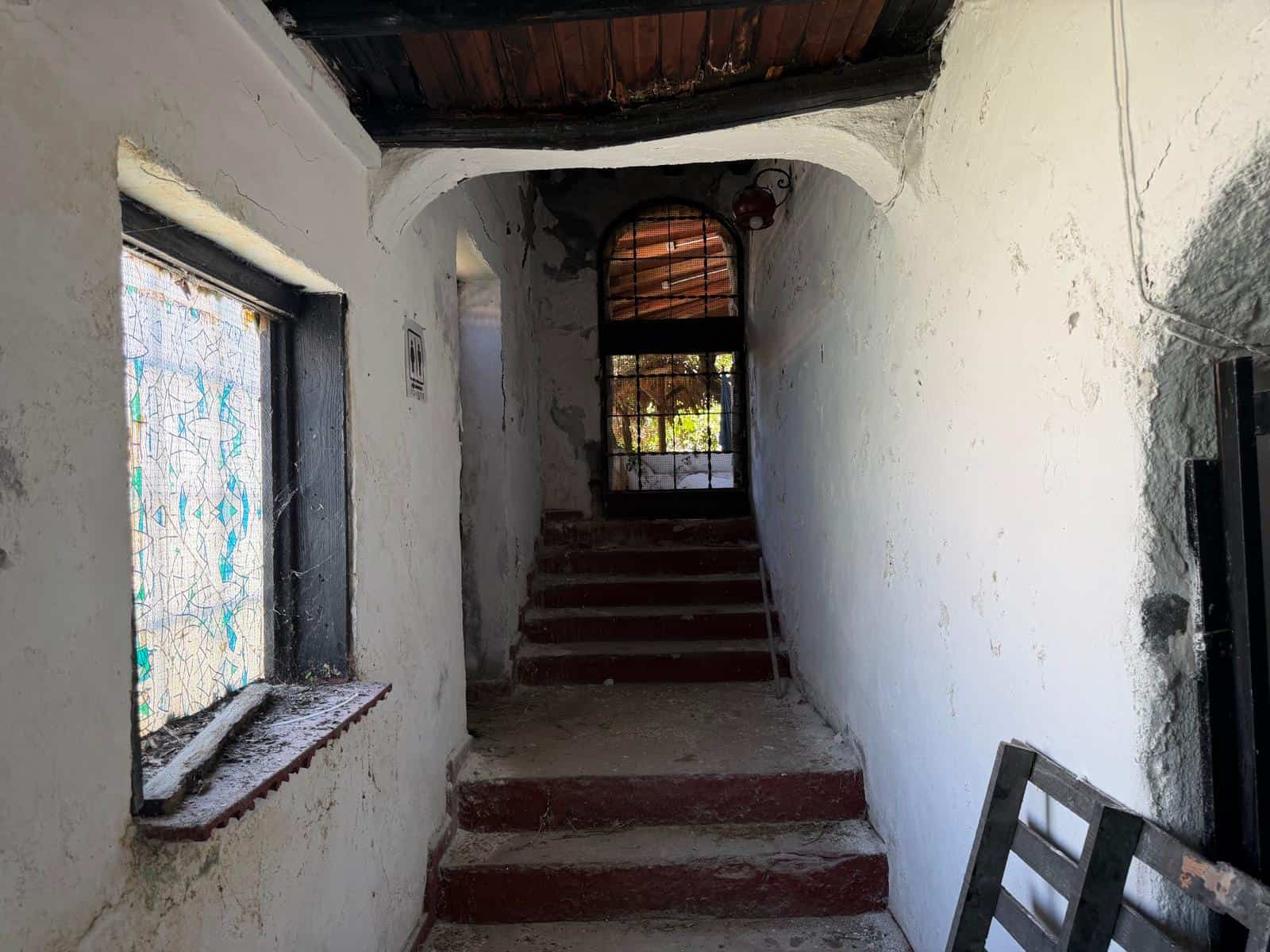 Scale interna di una casa rustica con pareti bianche, gradini in pietra e un arco in legno, illuminata dalla luce naturale che entra da una finestra e una porta sullo sfondo, creando un'atmosfera acco.