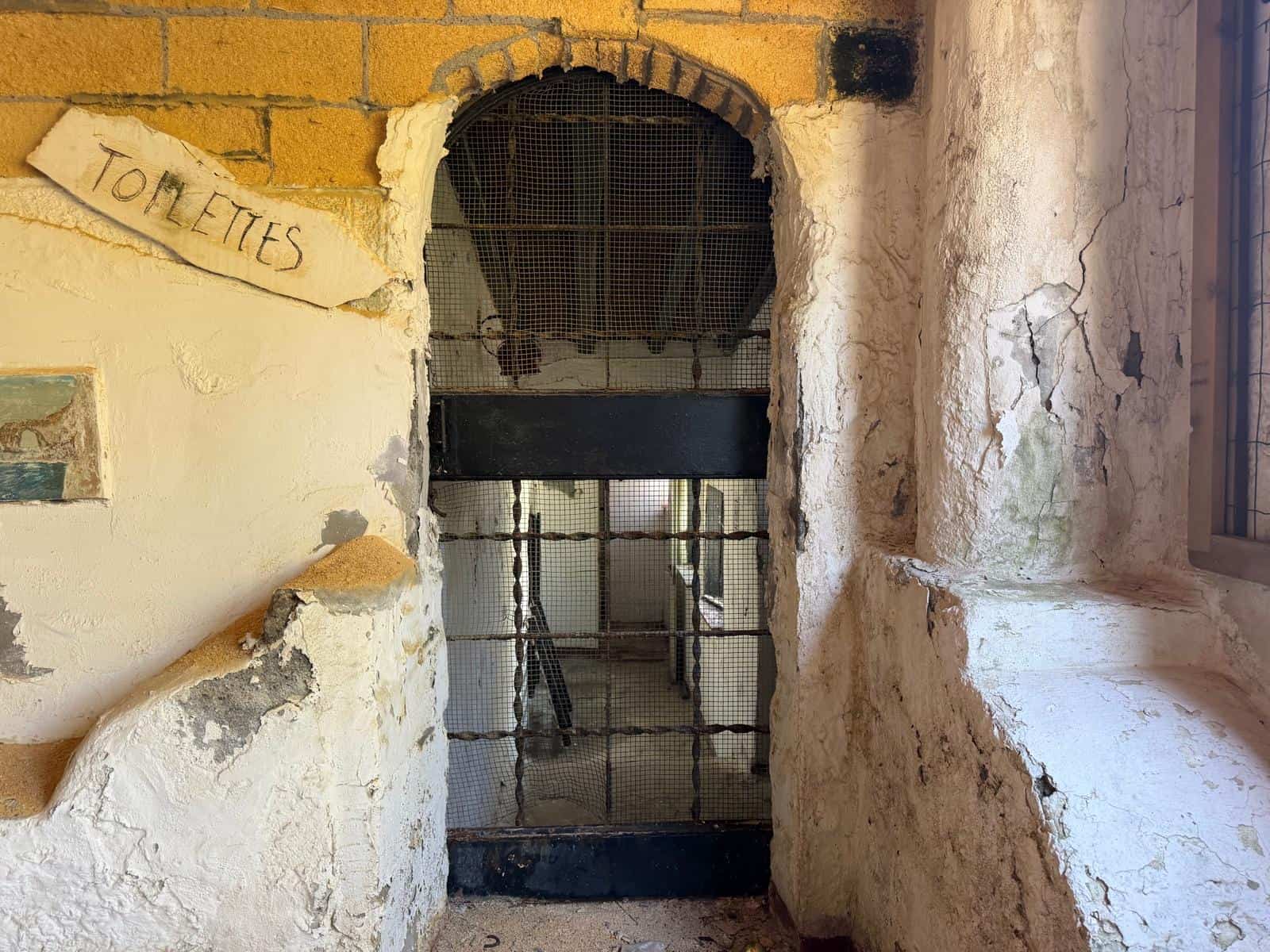 Un ingresso di un bagno pubblico in un edificio storico con pareti in pietra e segni di usura, con un cartello "TOILETTES" scritto a mano, e una grata di metallo che copre l'apertura.