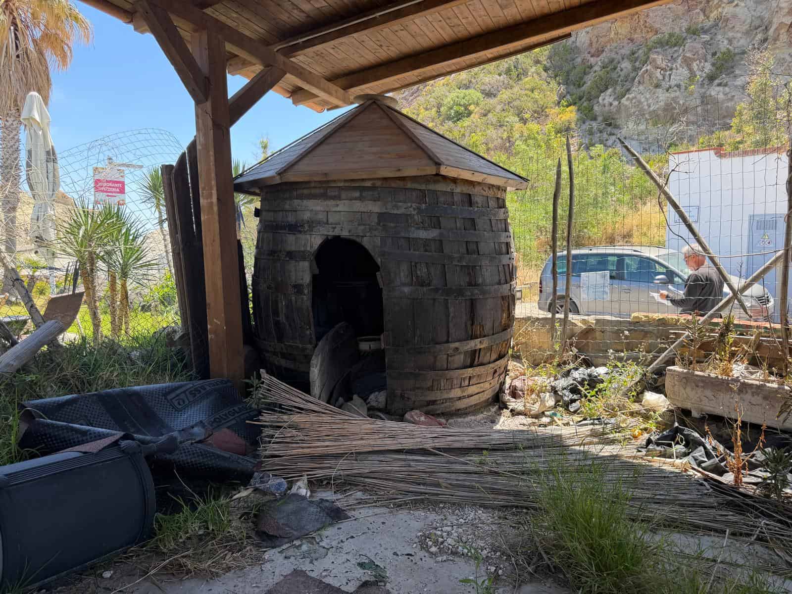 Casetta in legno rustica all'aperto, ambiente naturale con vegetazione e recinzione, sotto una tettoia in legno, con oggetti e attrezzi da giardino nelle vicinanze.